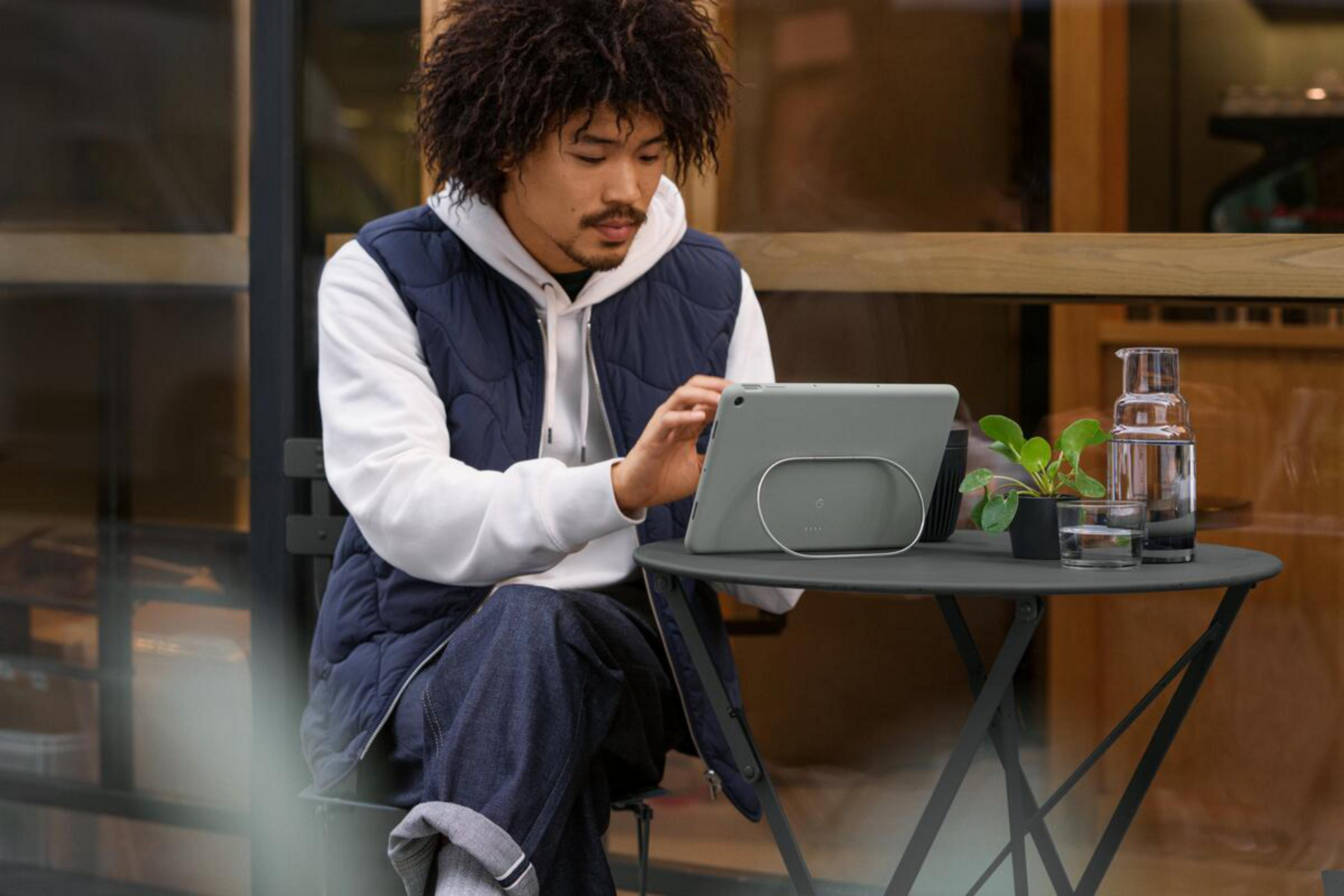 Mann sitzt draußen und benutzt ein Tablet. Ein Tisch mit Pflanze und Wasserglas ist in der Nähe.