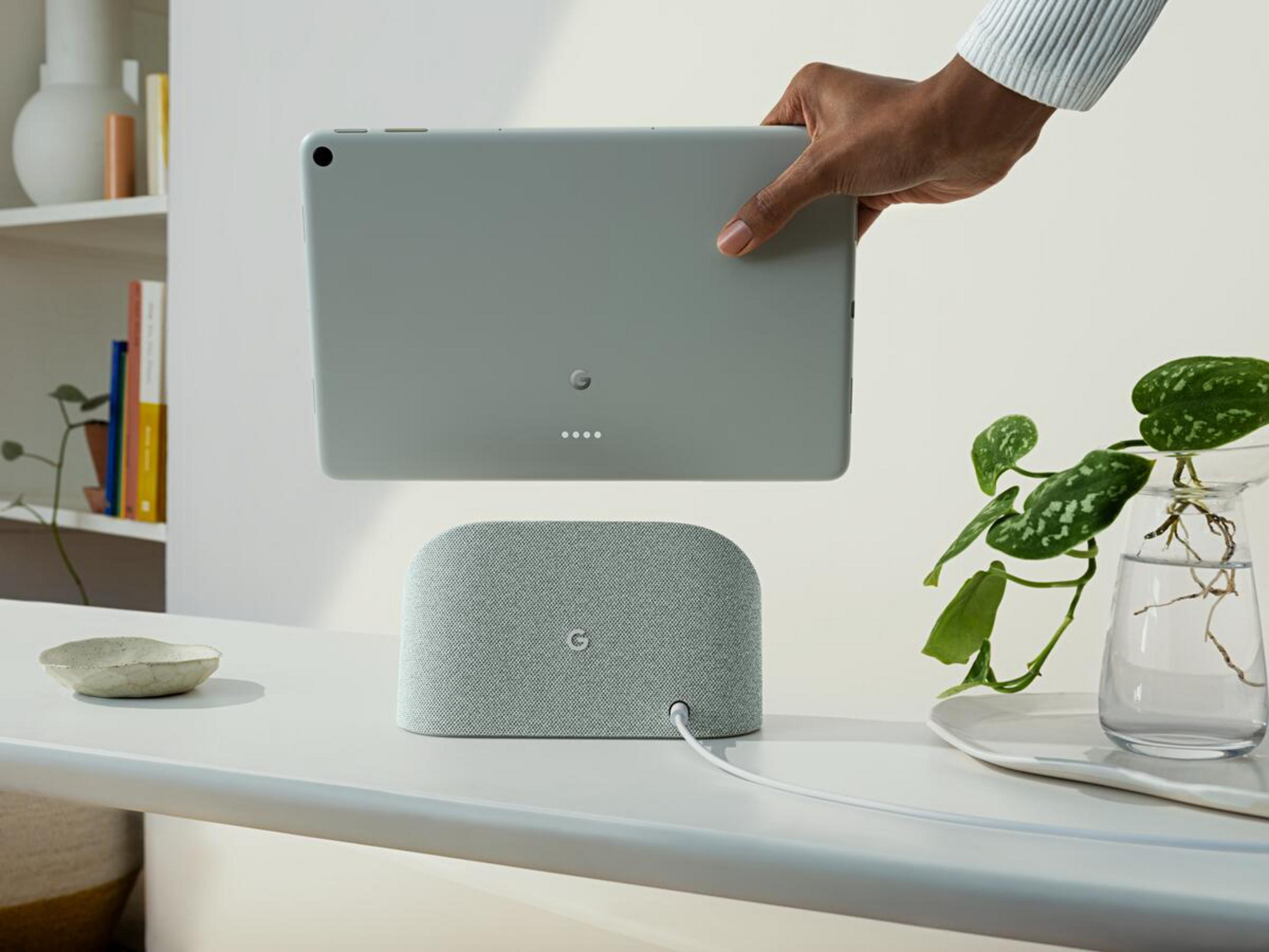 Hand hält graues Tablet über grauem Lautsprecher auf weißem Tisch. Pflanze sichtbar.