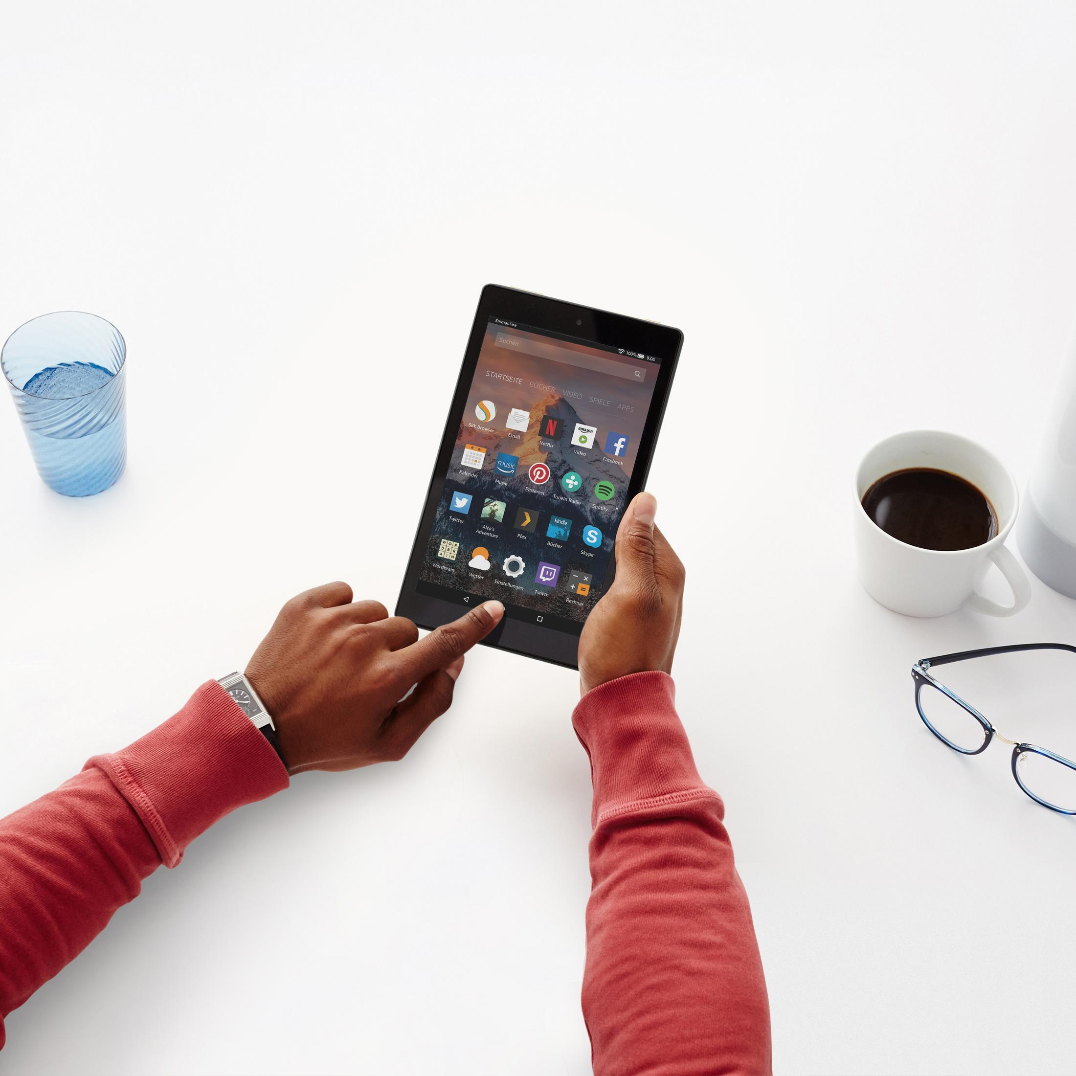 Hände halten ein schwarzes Tablet mit Touchscreen, mit einer Tasse Kaffee, Brille und einem blauen Glas auf weißer Oberfläche.