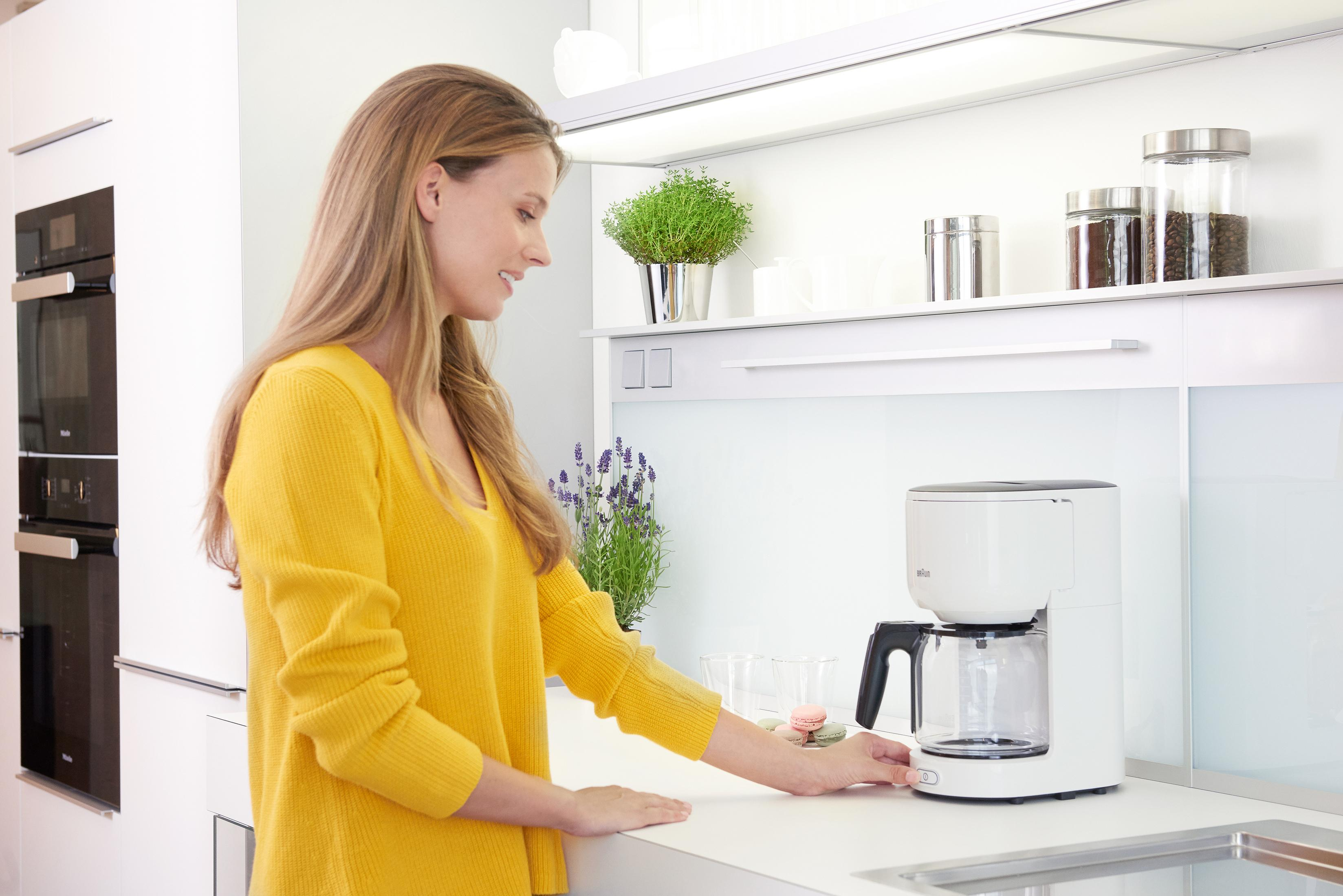 Kobieta w żółtym swetrze używa ekspresu do kawy. Kuchnia z półkami, roślinami w tle.