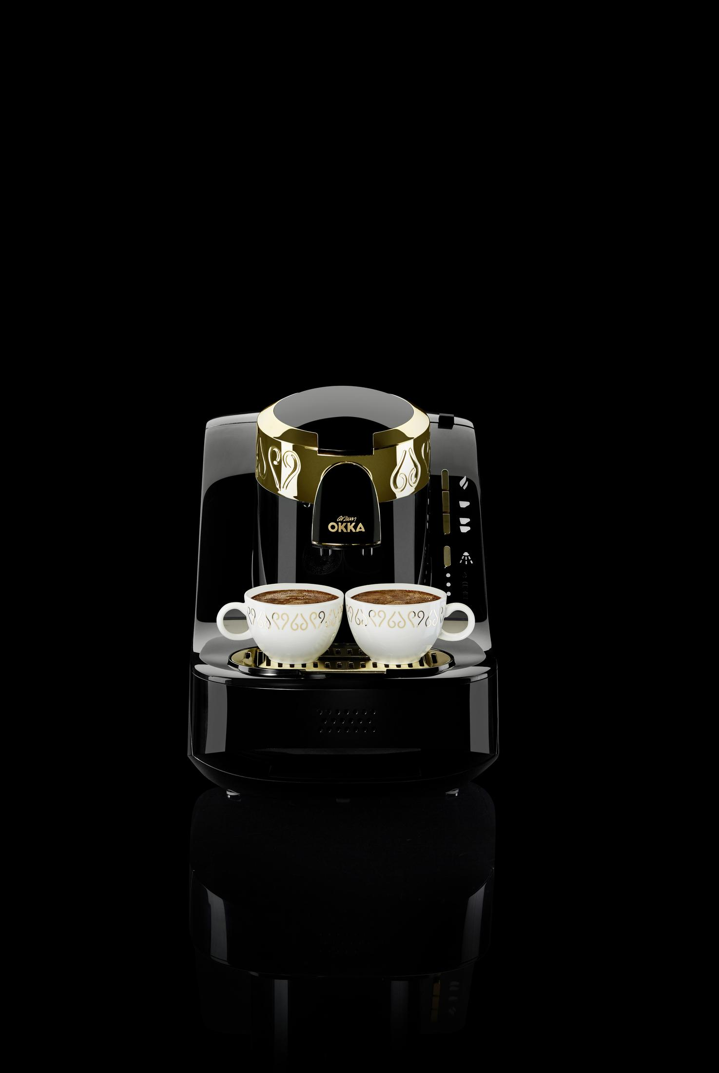Caffettiera nera e oro con due tazze bianche piene di caffè, su una superficie nera.