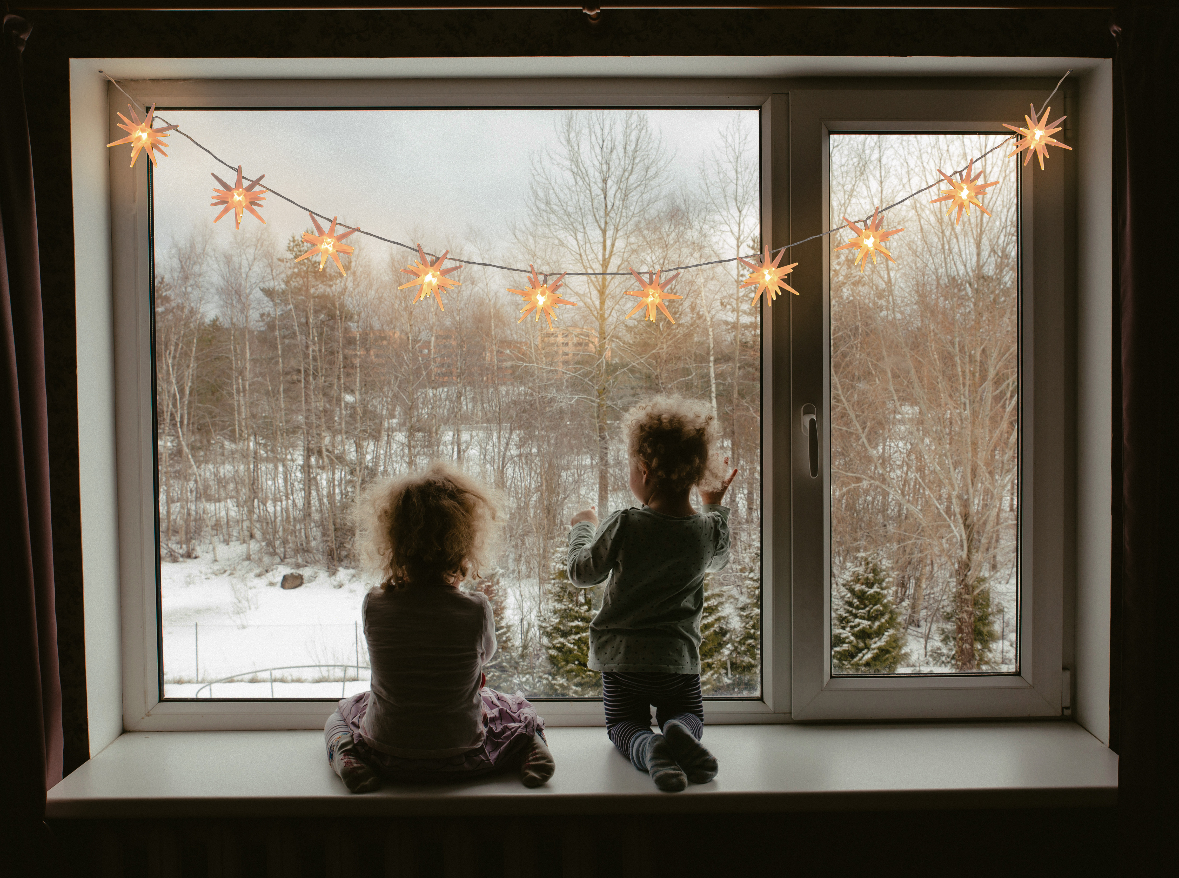 Zwei Kinder schauen aus einem Fenster mit sternförmigen Lichtern. Schneelandschaft draußen sichtbar.