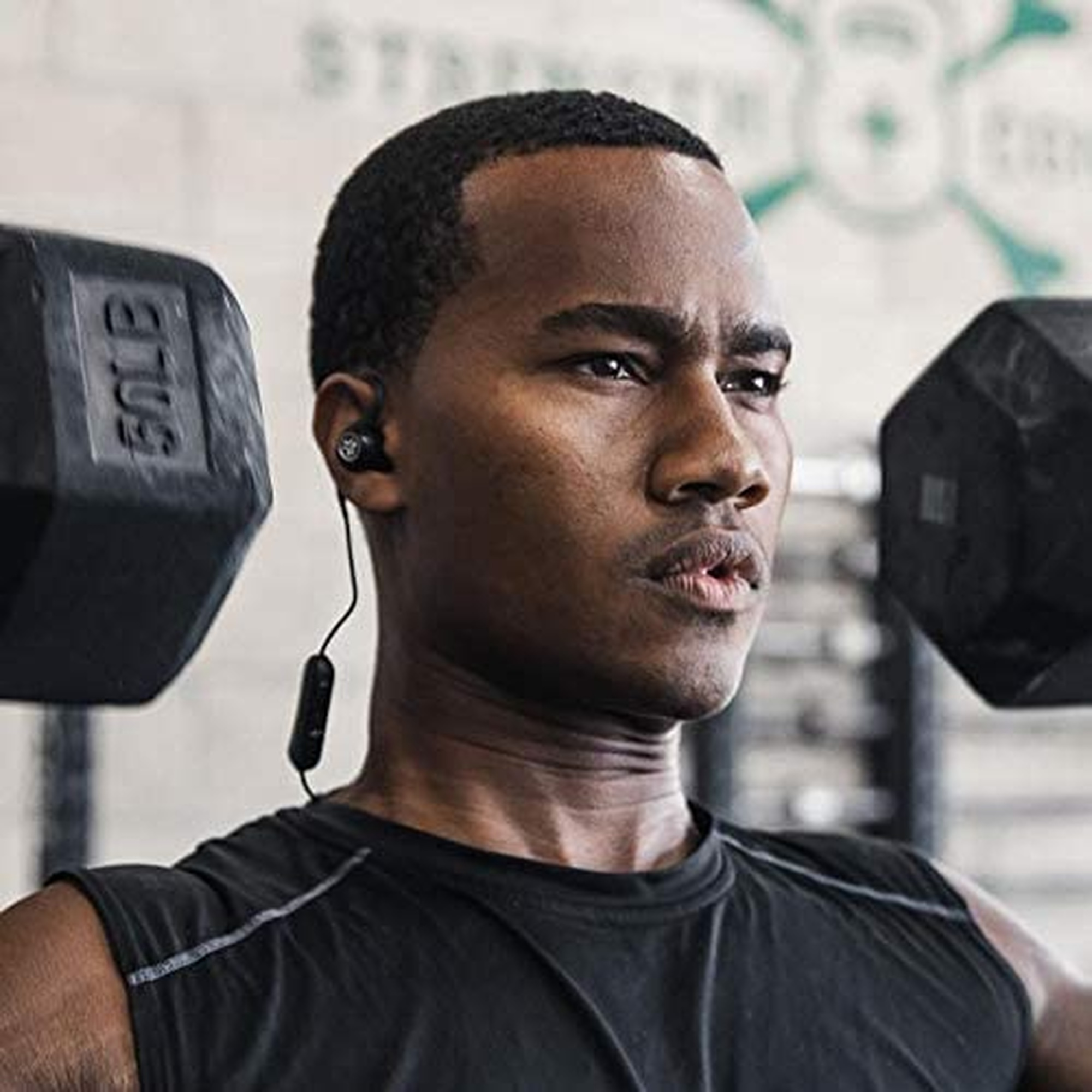 Czarny mężczyzna noszący słuchawki podczas ćwiczeń. Ma poważny wyraz twarzy i trzyma ciężarki.