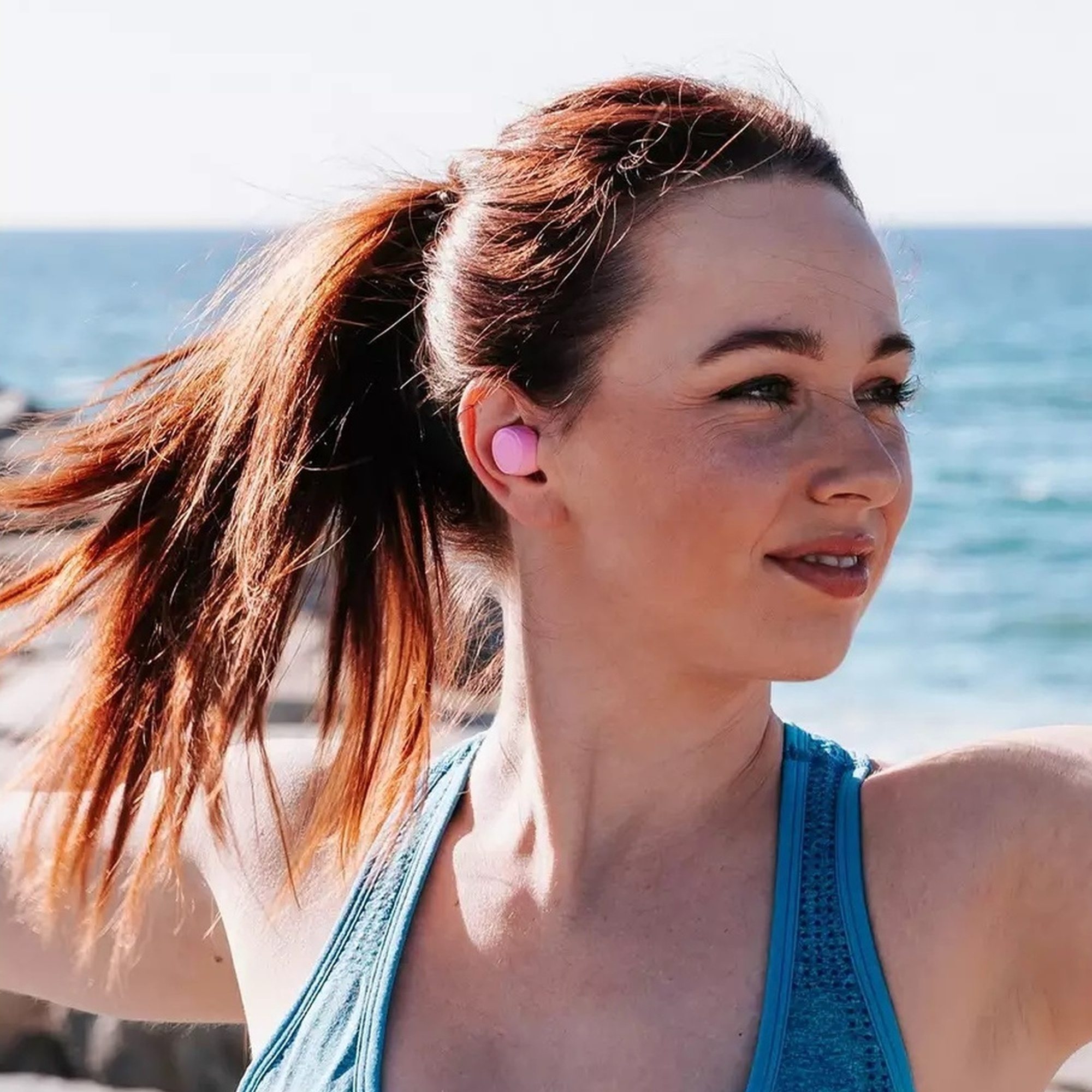 Kobieta z rudymi włosami w różowych słuchawkach, uśmiechnięta. Plaża i morze w tle.