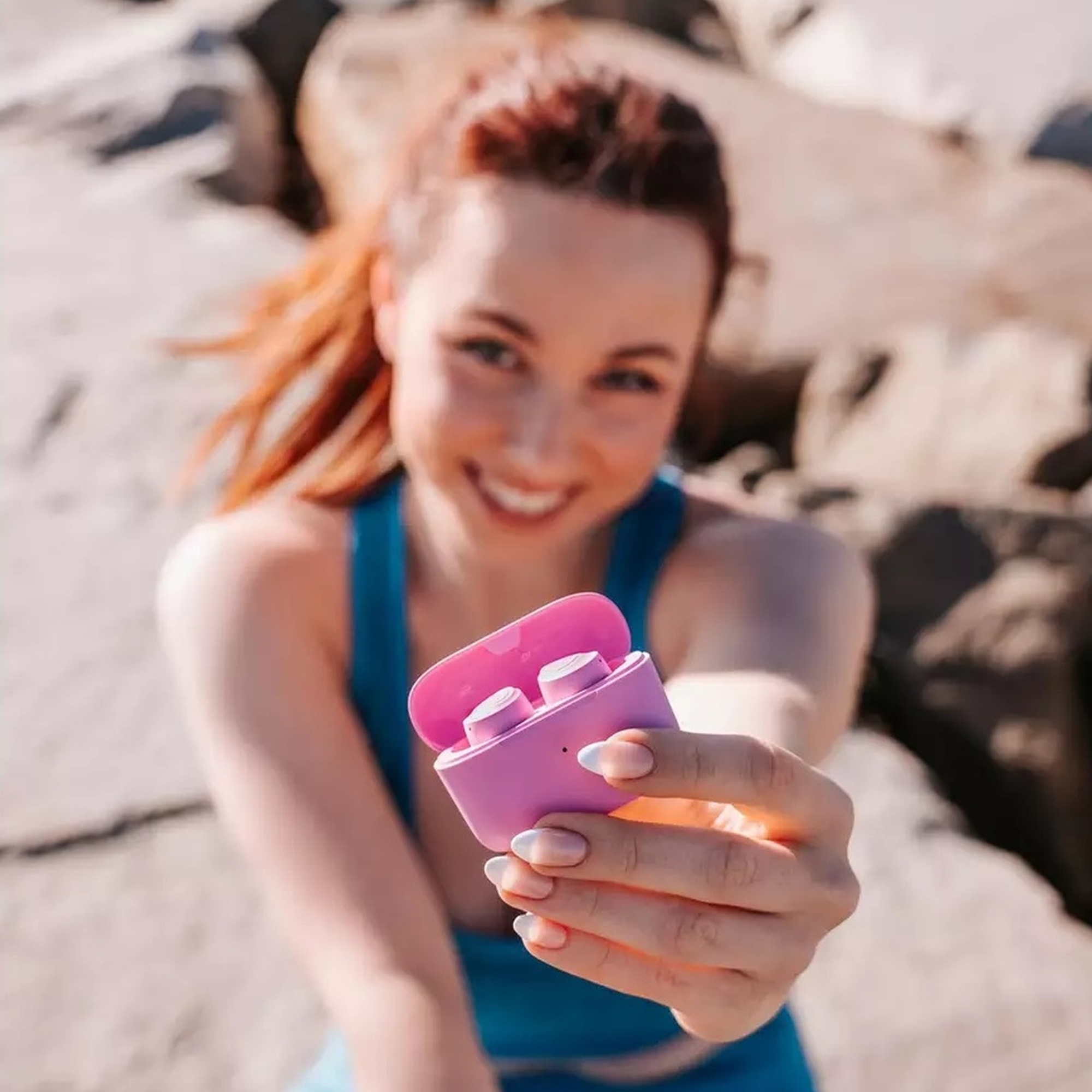 Uśmiechnięta kobieta trzymająca różowe etui ze słuchawkami. Na zewnątrz, blisko skał.