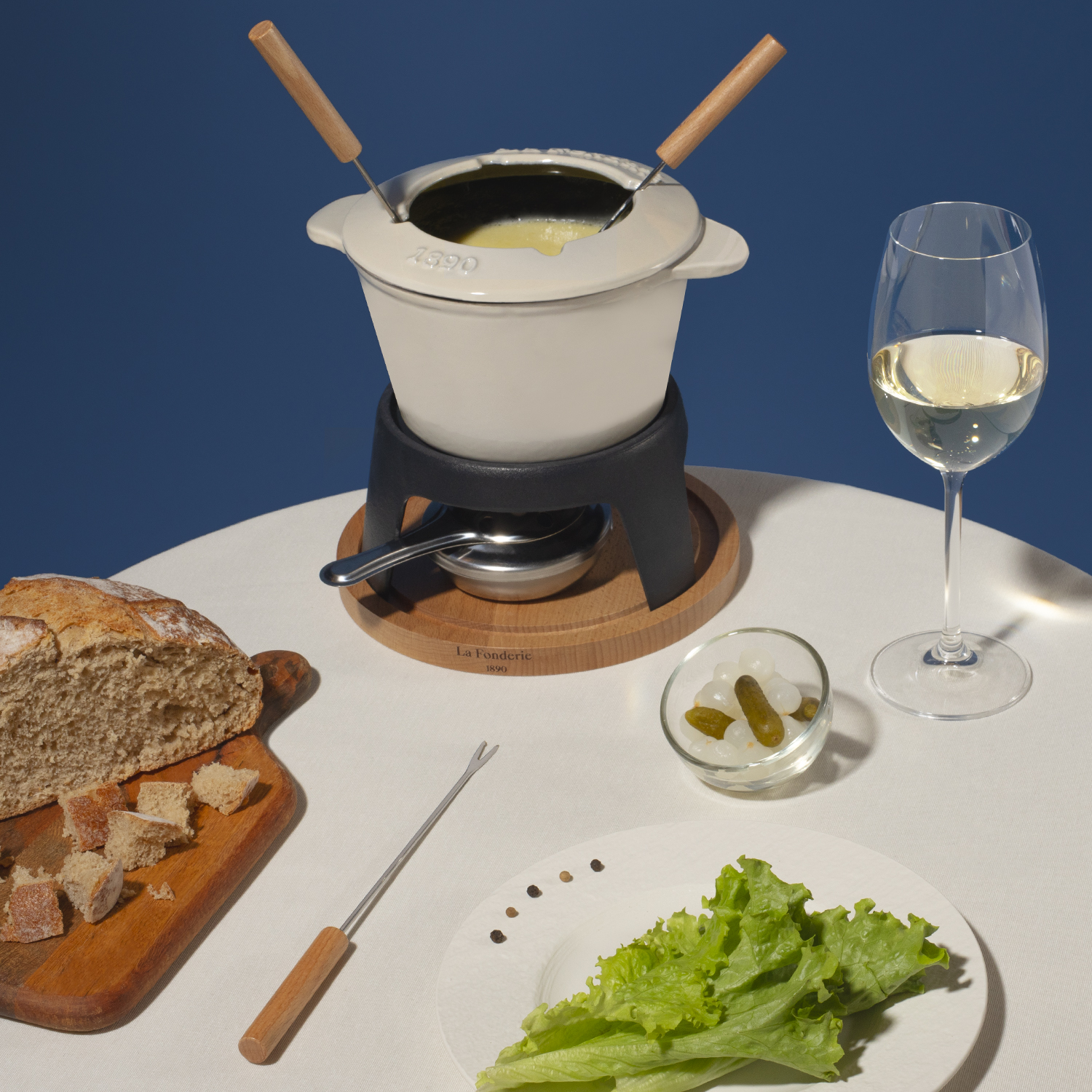 Ein Fondue-Set mit Käse, Brot, Essiggurken und einem Glas Weißwein steht auf einem Tisch.