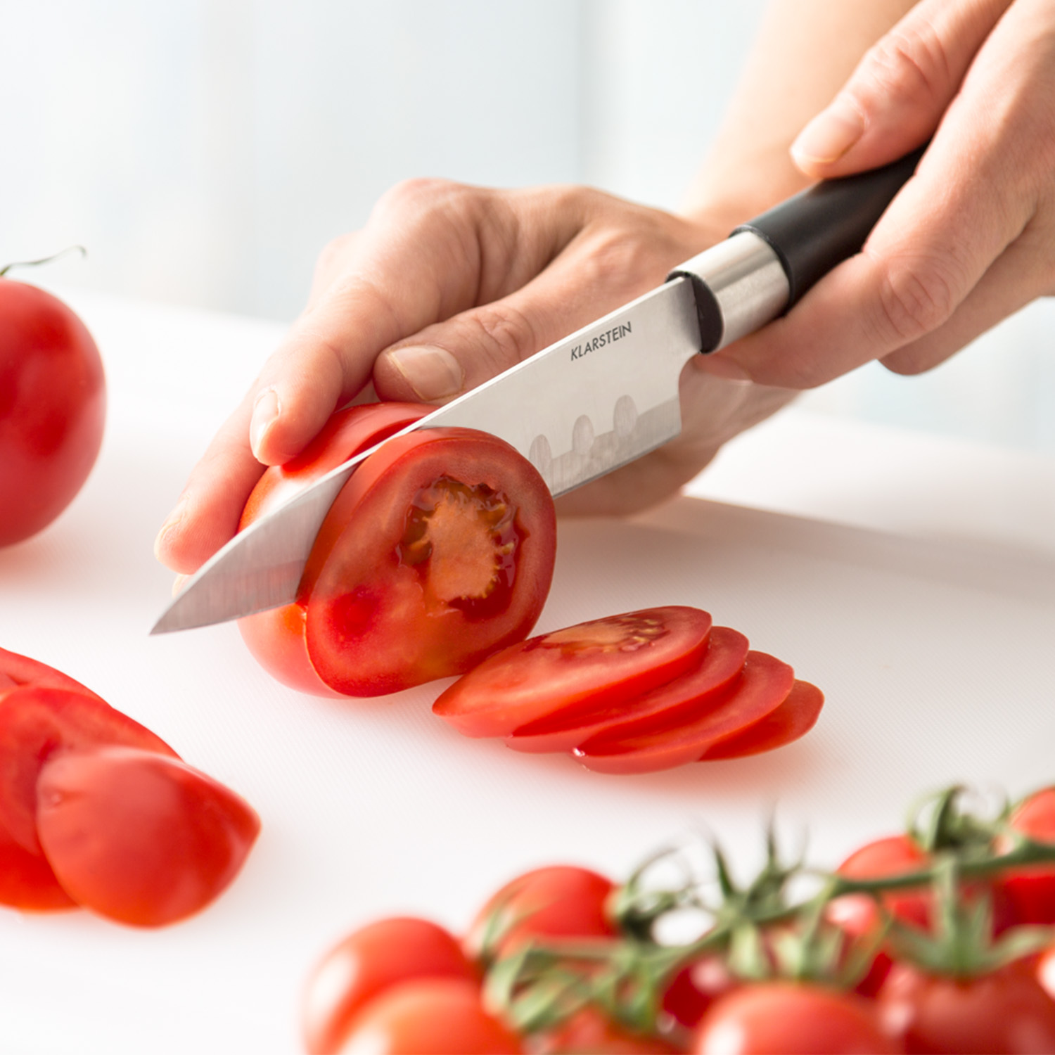 Eine Hand schneidet eine Tomate auf einem weißen Schneidebrett mit einem Messer.