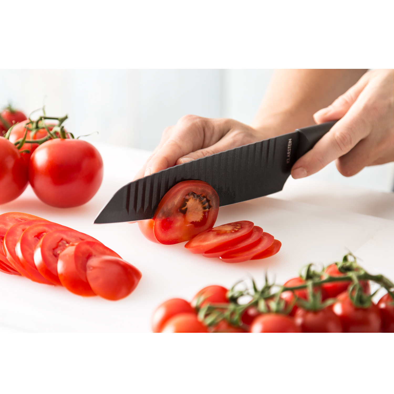 Person schneidet Tomate mit schwarzem Messer auf weißem Schneidebrett. Geschnittene Tomaten sind vorhanden.
