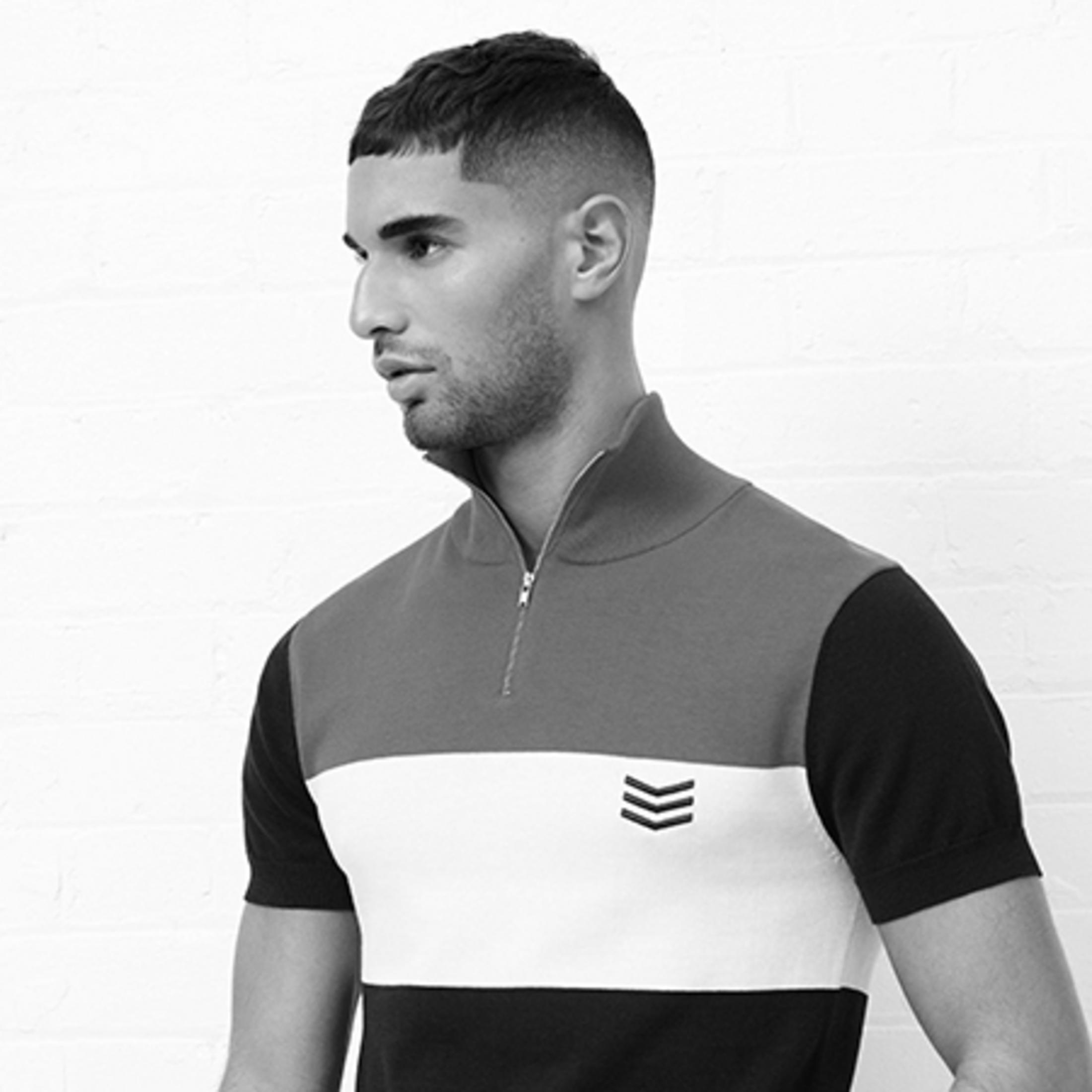 Mężczyzna w czarno-biało-szarej koszuli, patrzący na bok, na tle białej ceglanej ściany.