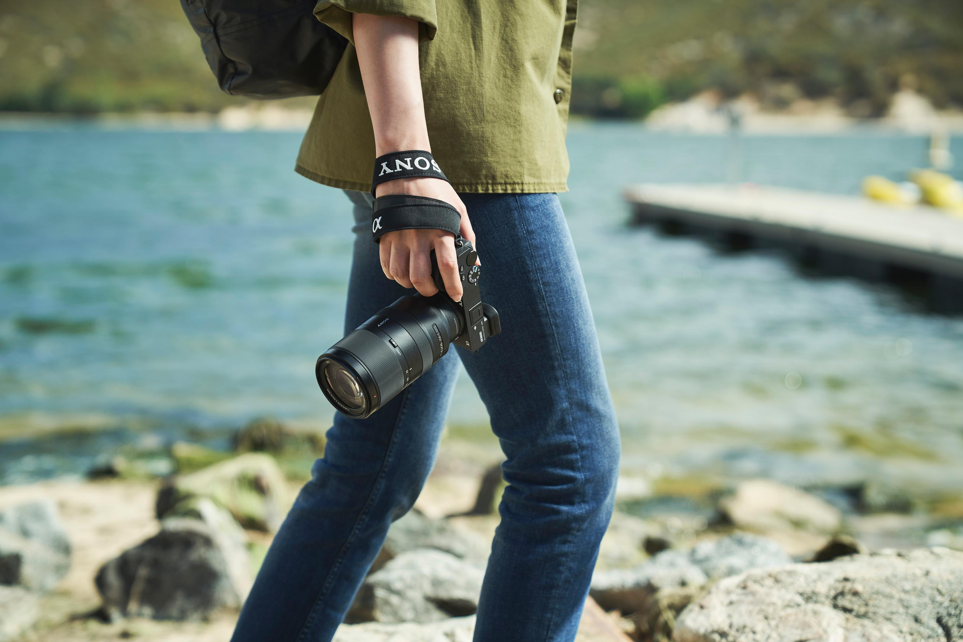 Osoba idzie wzdłuż jeziora trzymając aparat. Ma na sobie dżinsy i zieloną koszulę.