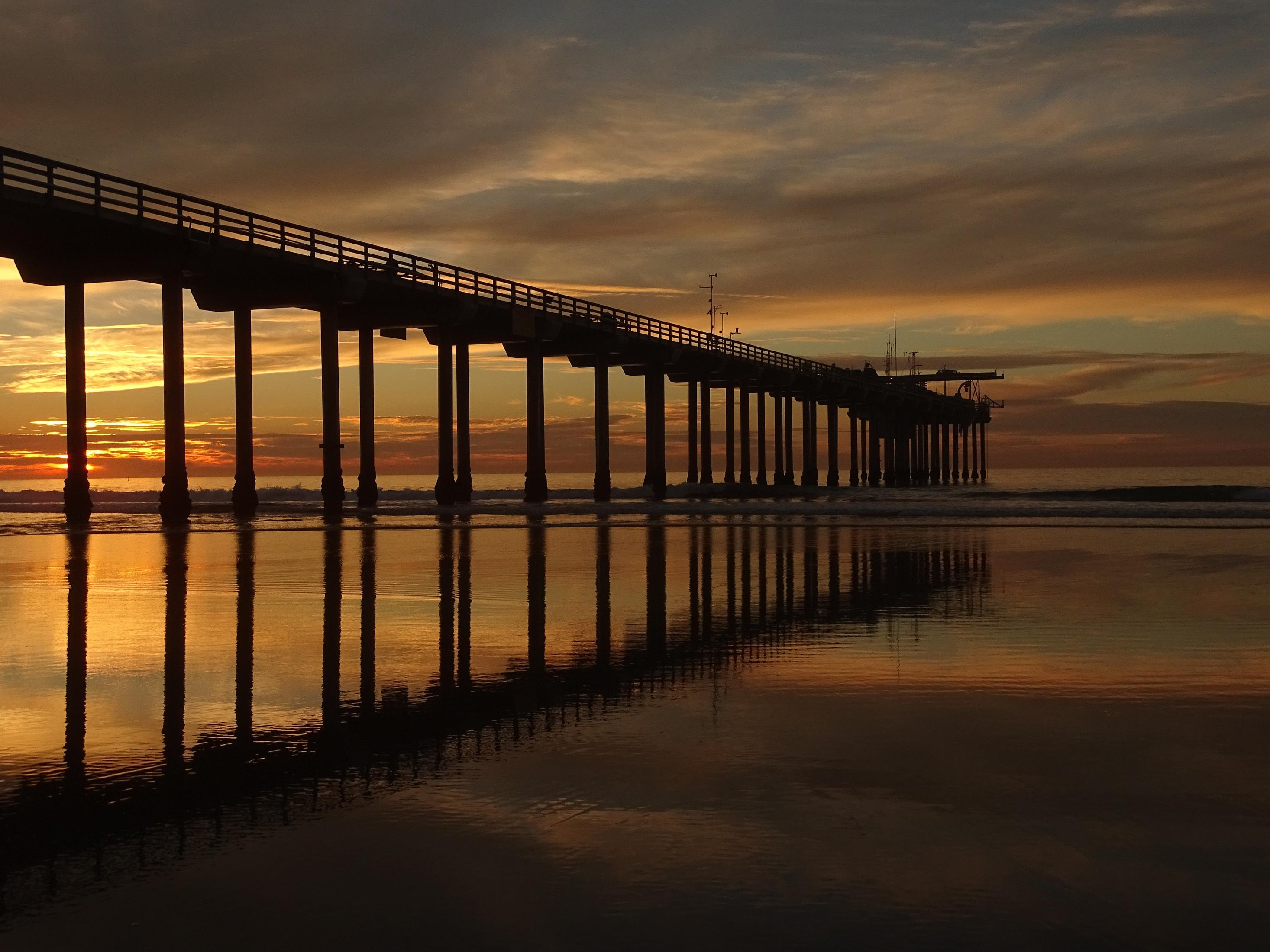 Pier bei Sonnenuntergang, spiegelt sich im nassen Sand. Orange und dunkelblaue Farben.