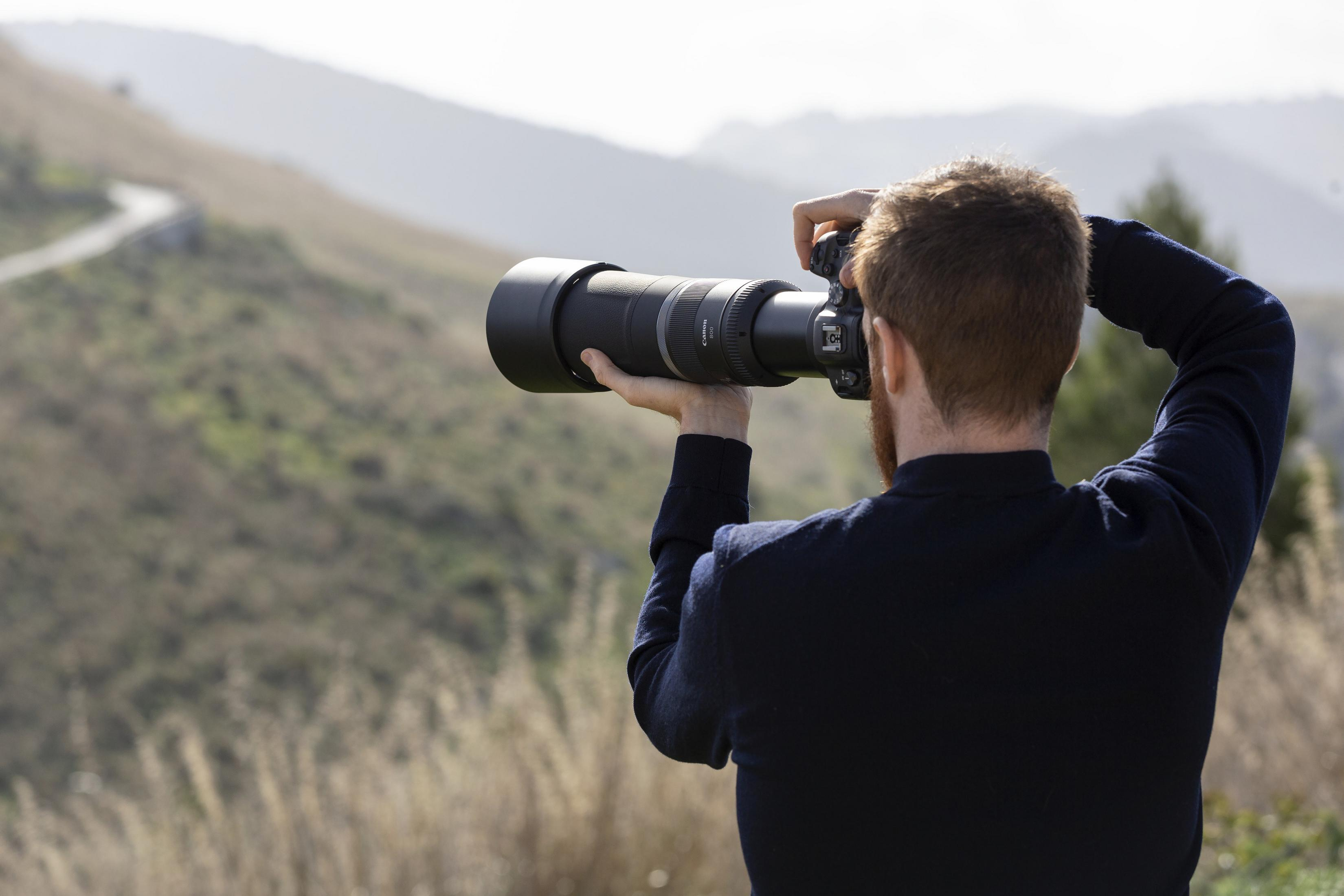 Photographer outdoors, holding a camera with a large lens. Natural setting, blurry background.
