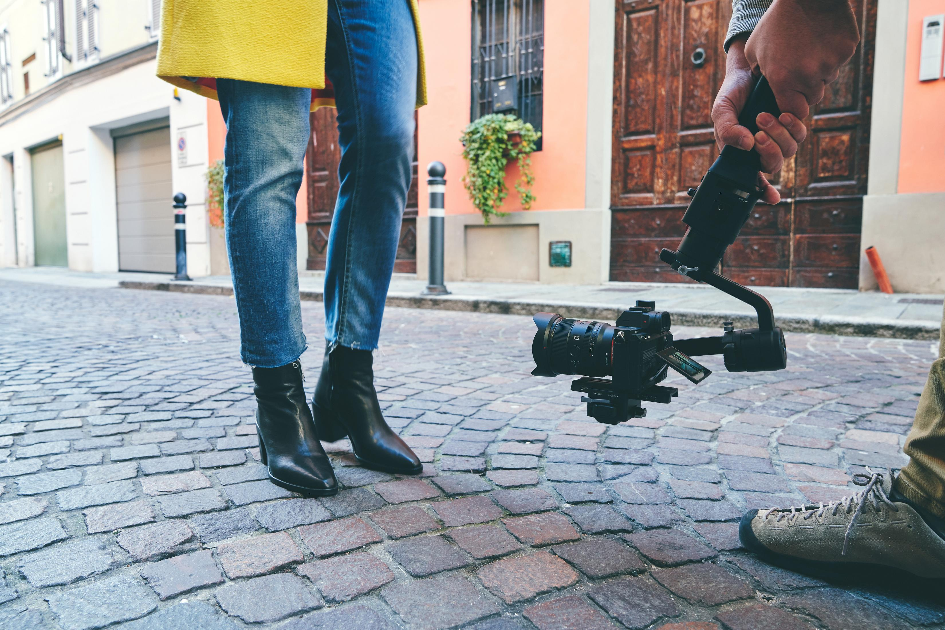 Osoba trzymająca kamerę na gimbalu, filmująca kobietę stojącą na brukowanej ulicy.