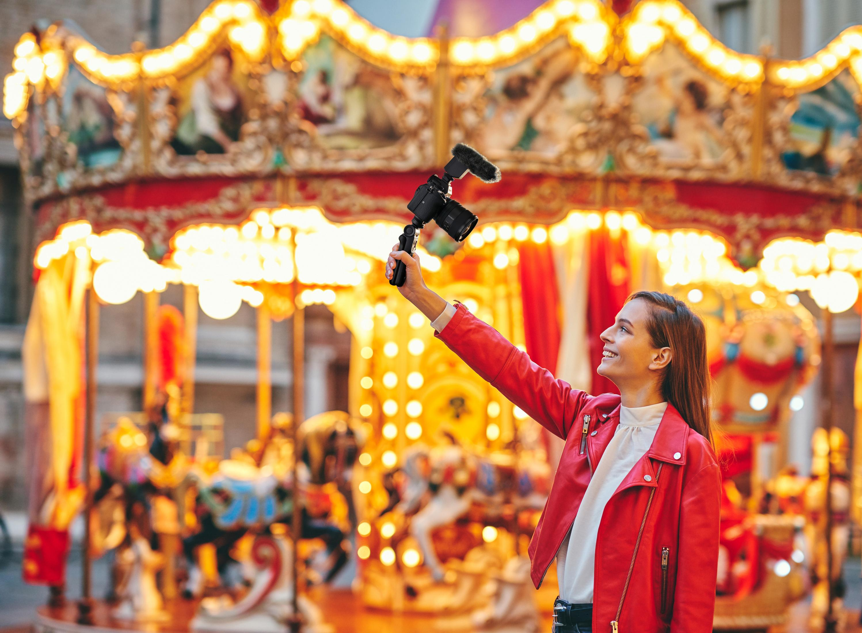Kobieta w czerwonej kurtce z aparatem uśmiecha się w pobliżu karuzeli ze światłami. Tło jest rozmyte.