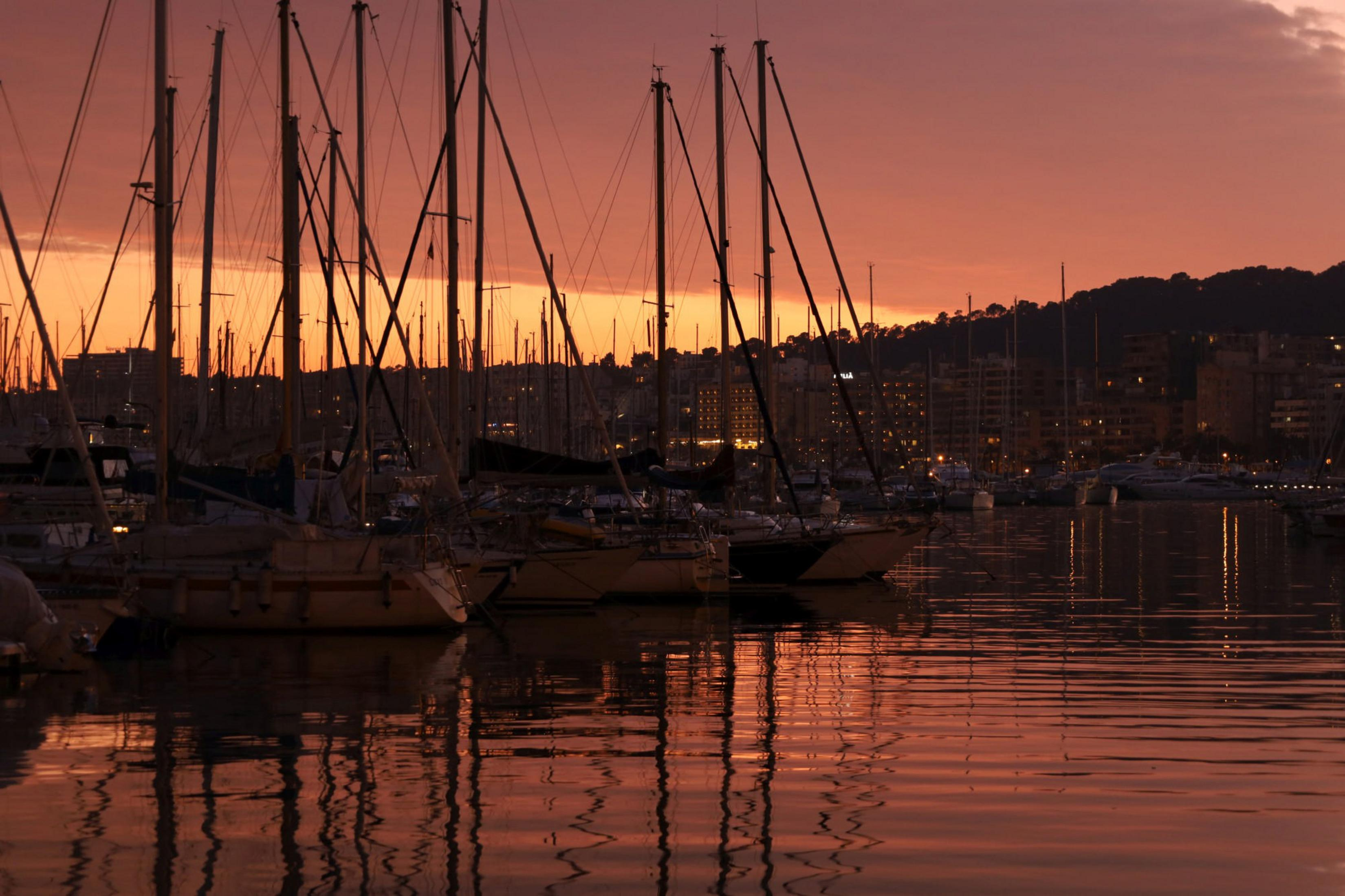 Żaglówki w porcie o zachodzie słońca, z odbiciami w wodzie i budynkami w tle.