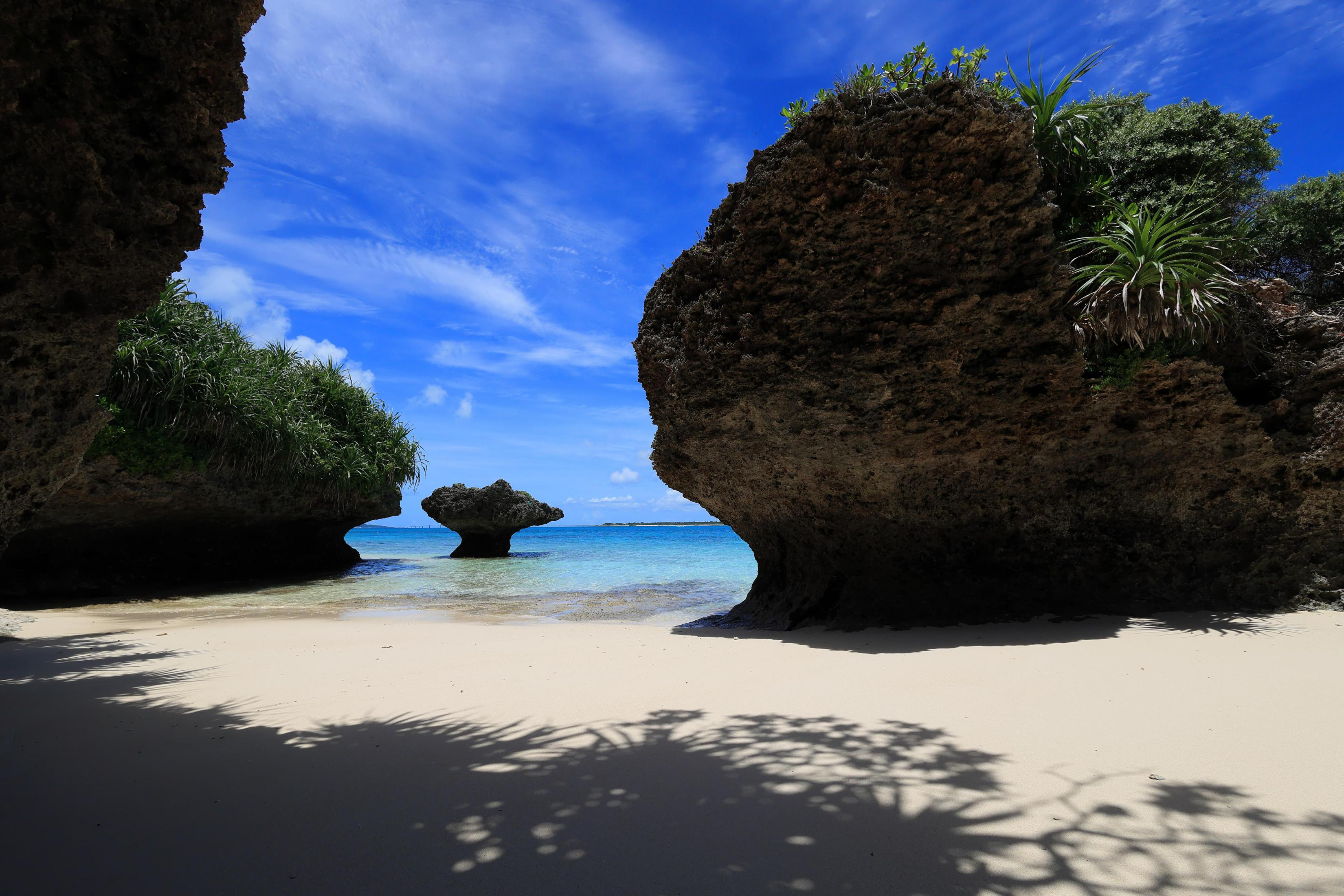 Beach scene with white sand, blue water, and rock formations. Sky is clear blue.