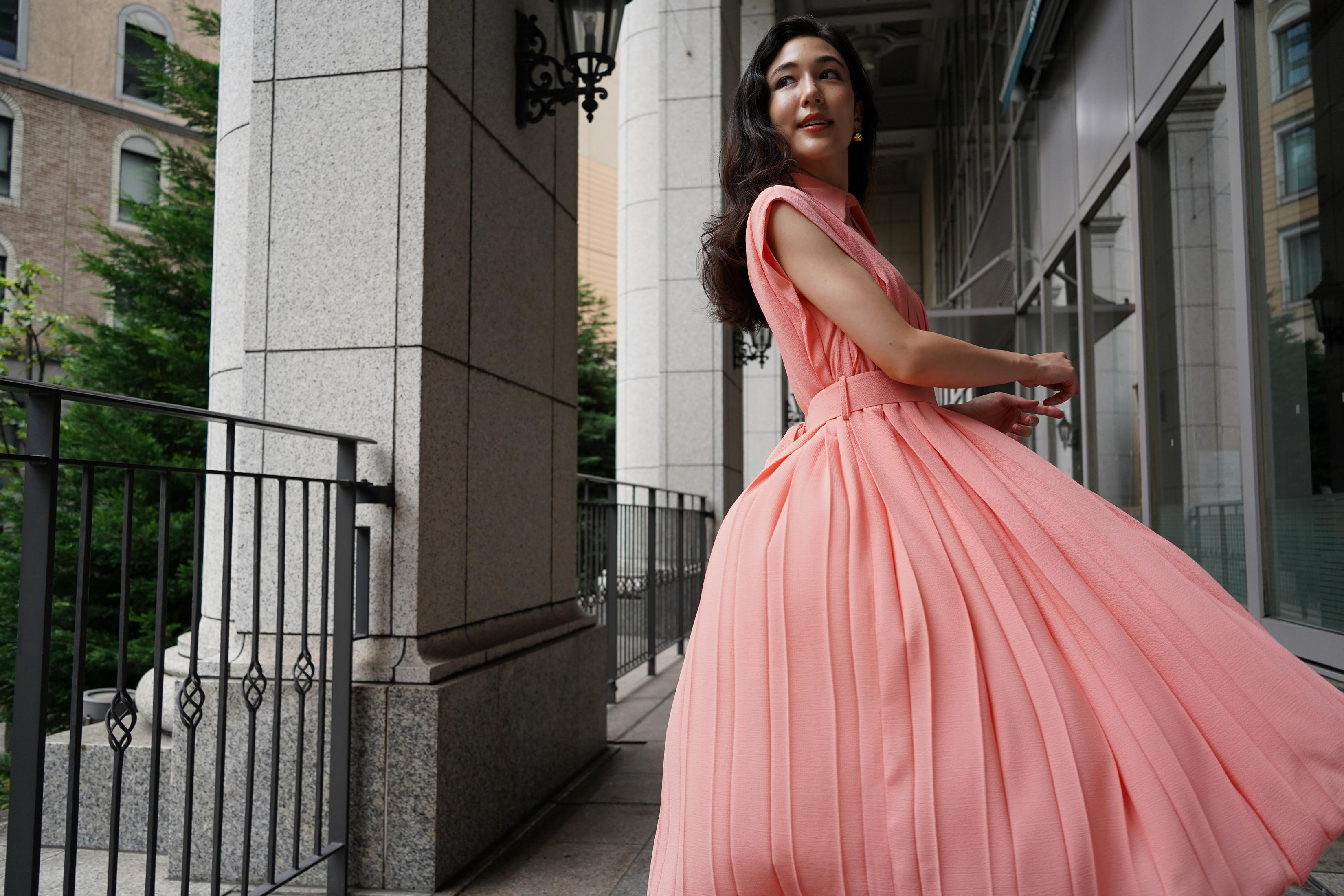 Kobieta w różowej sukience kręci się na zewnątrz budynku z kamiennymi kolumnami i metalową balustradą.