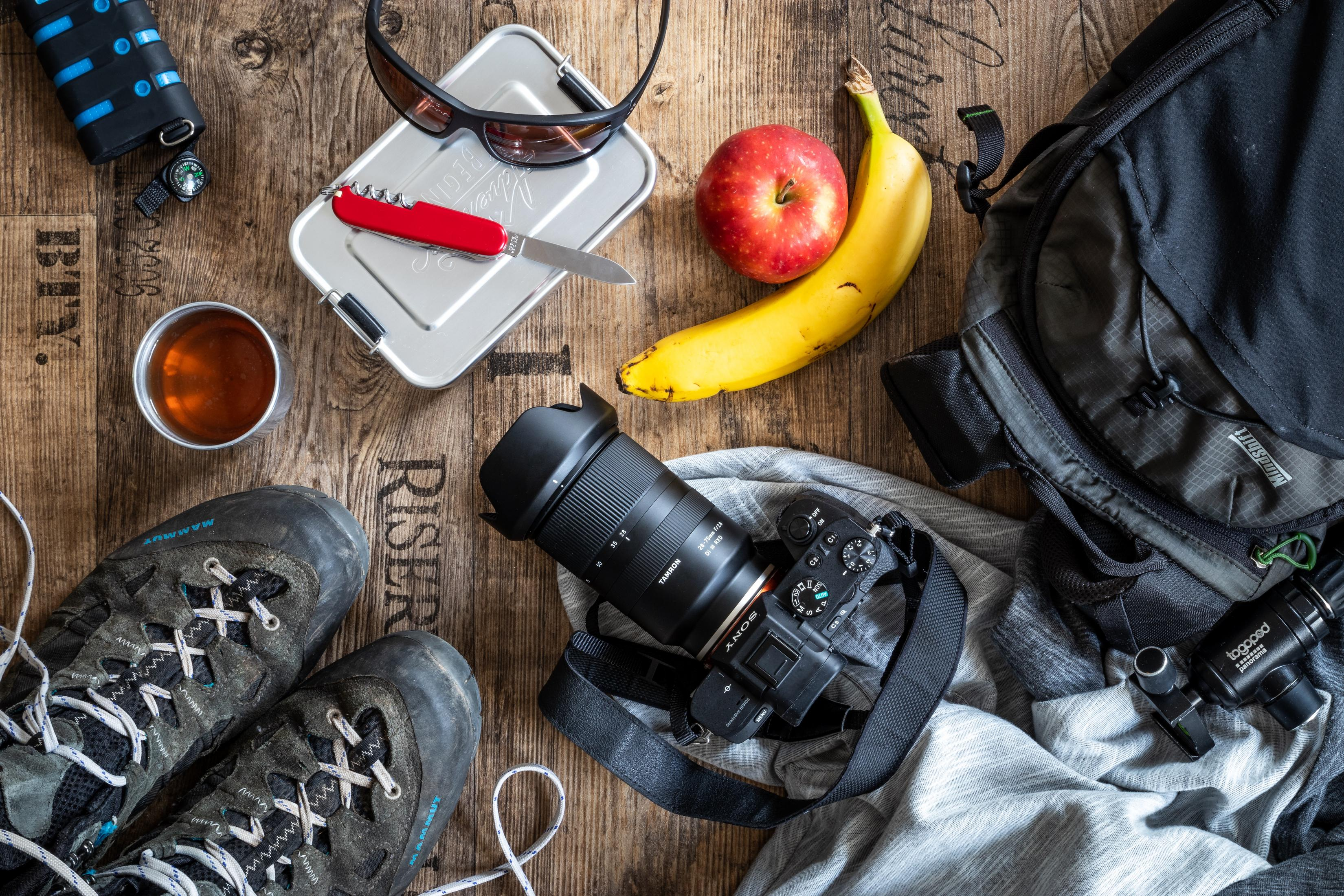 Draufsicht auf ein Picknick-Setup mit Essen, Kamera und Wanderausrüstung auf einer Holzoberfläche.