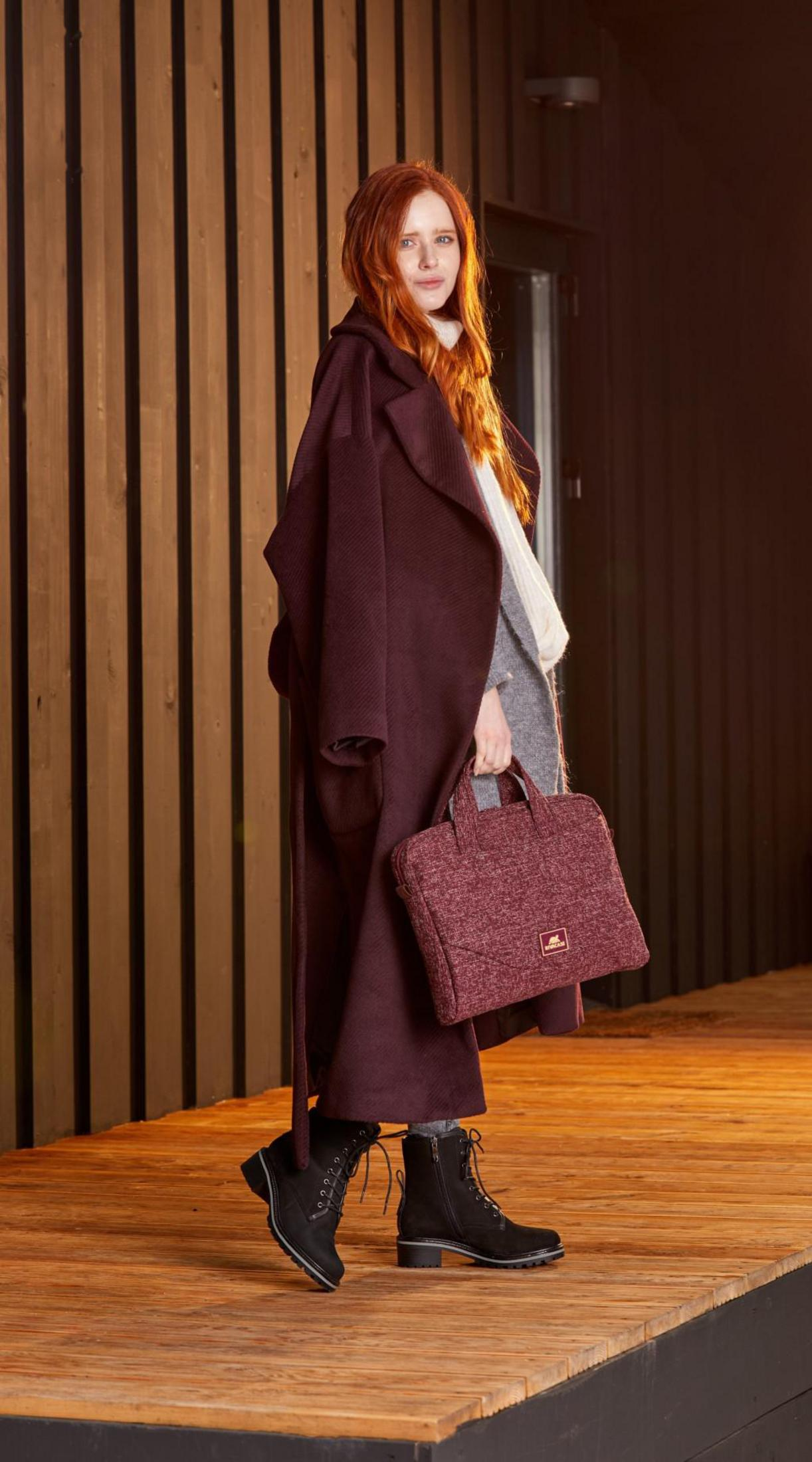 Kobieta w bordowym płaszczu i butach, z torbą, stoi na drewnianej powierzchni.