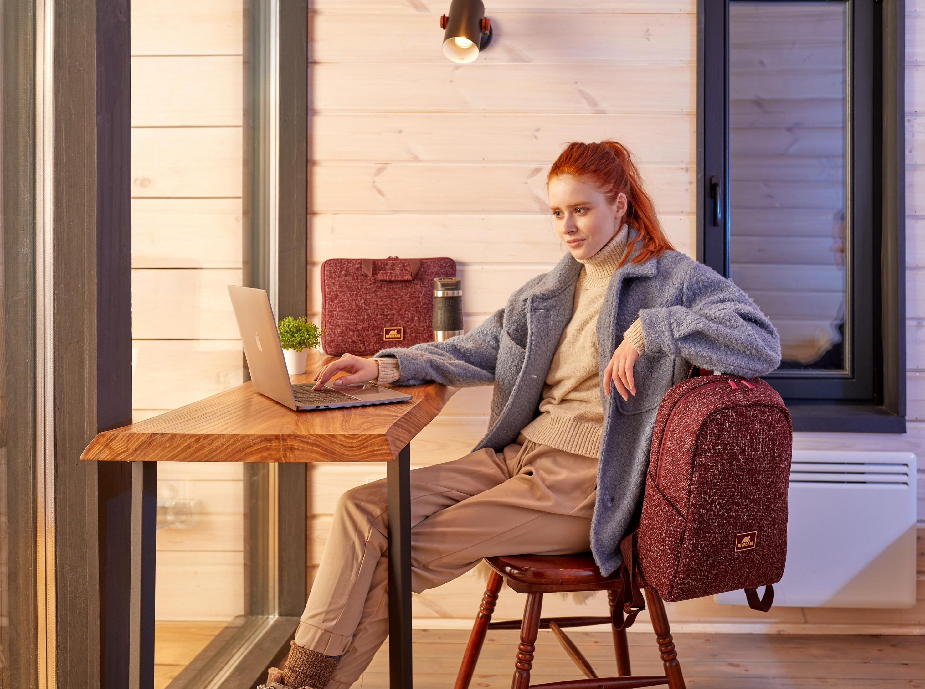 Kobieta siedzi przy drewnianym biurku z laptopem. Widoczny plecak i pokrowiec na laptopa.