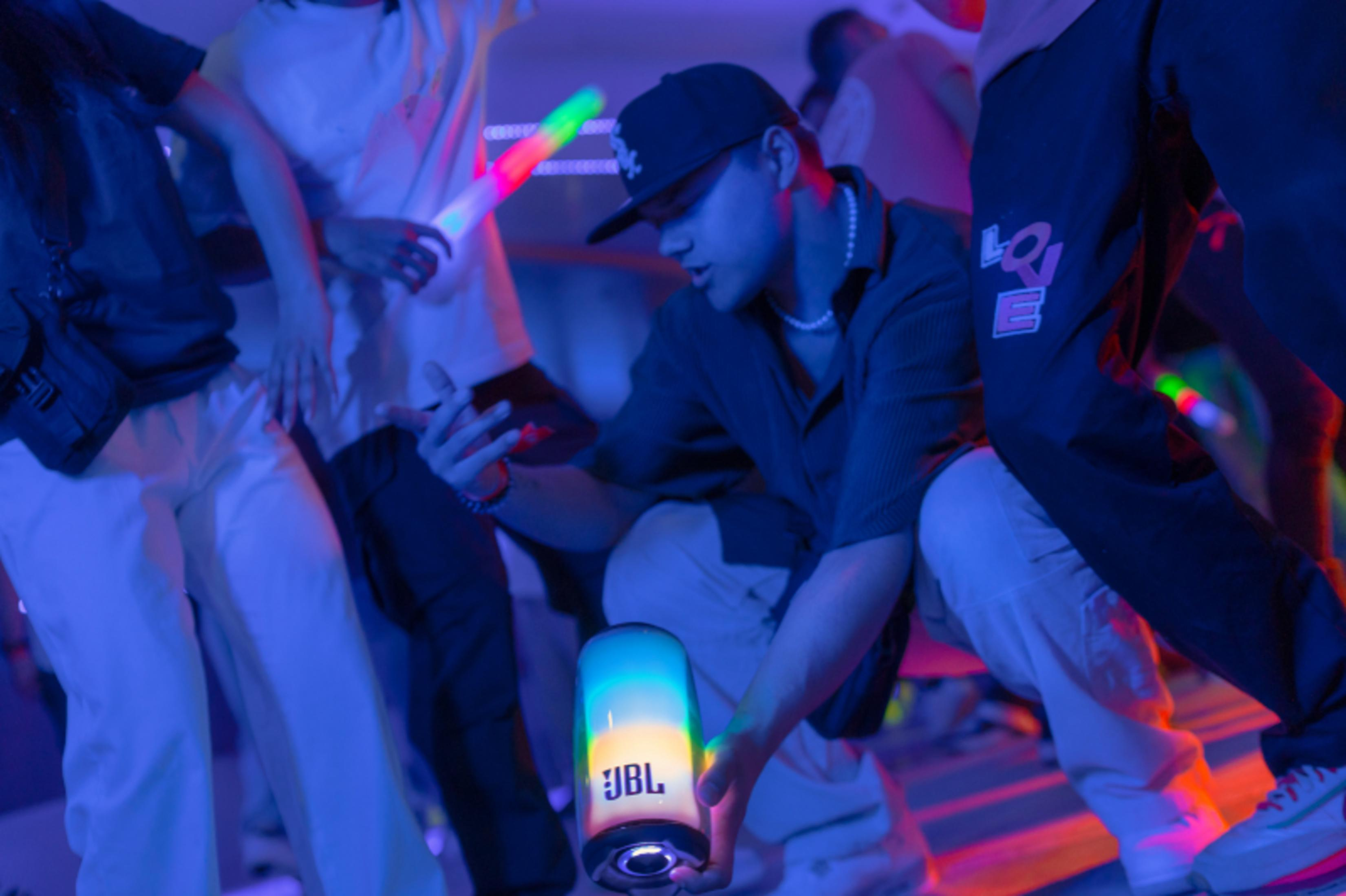Leute tanzen in einem Club, ein Mann hält einen JBL-Lautsprecher. Bunte Lichteffekte beleuchten die Szene.