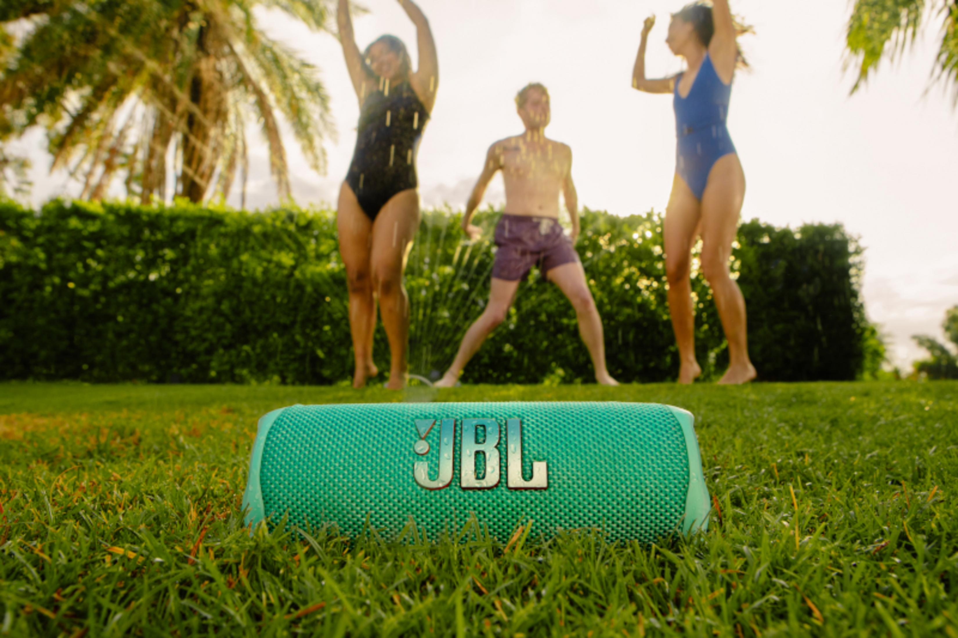 Altavoz JBL en la hierba con tres personas en trajes de baño jugando en el fondo.
