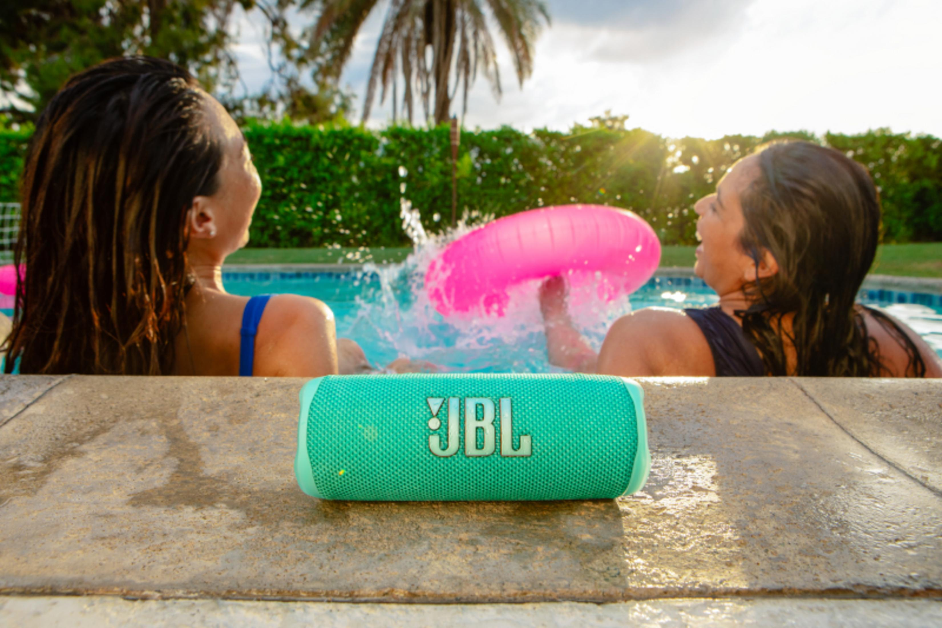 Dos mujeres en una piscina junto a un altavoz JBL verde. Una sostiene un anillo inflable rosa.