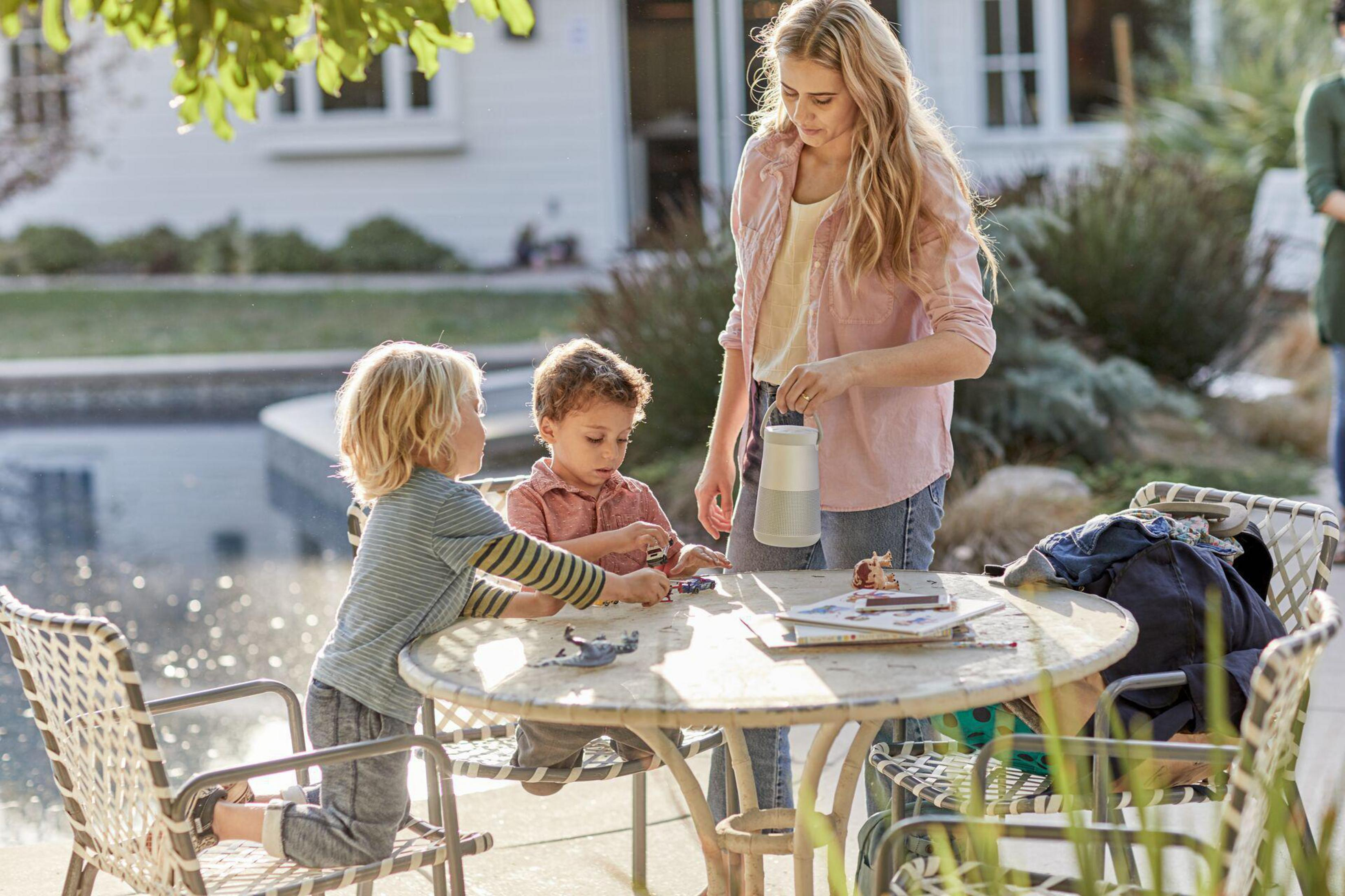 Frau und zwei Kinder an einem Tisch, spielen mit Spielzeug und einer Wasserflasche im Garten.