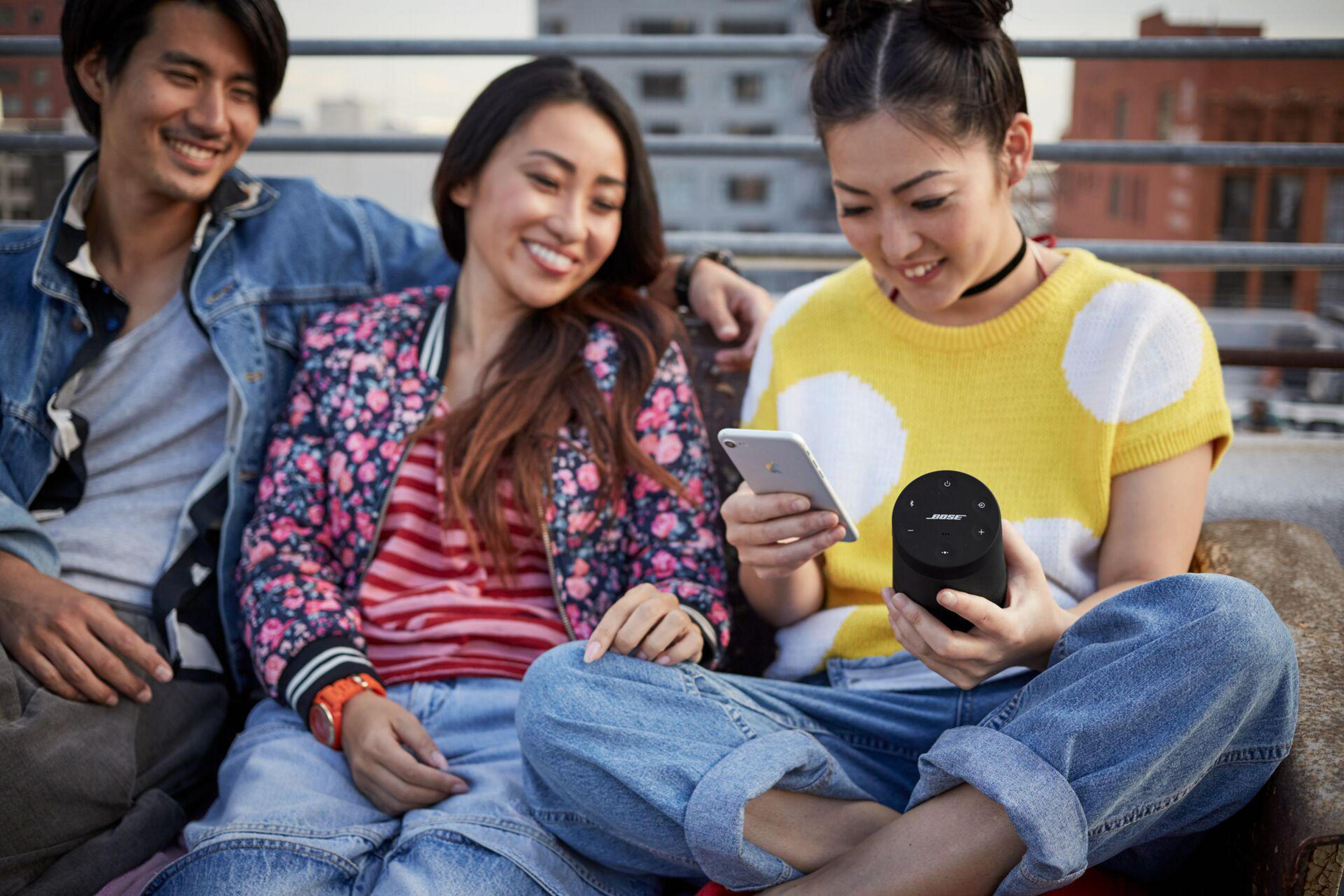 Tre persone sono sedute insieme, guardando un telefono e un altoparlante. Lo sfondo è un tetto.