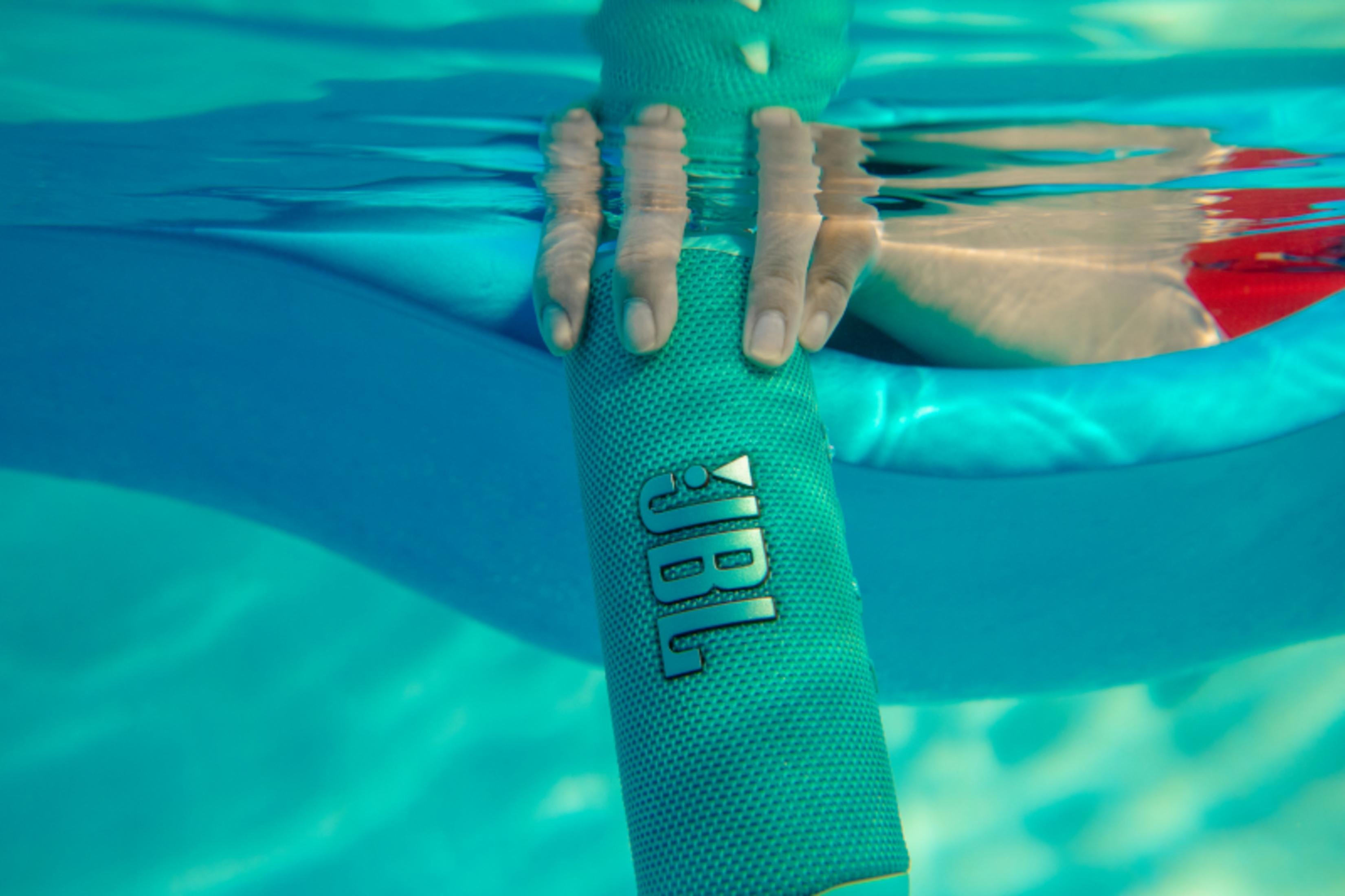 Una mano sosteniendo un altavoz JBL turquesa bajo el agua, con una piscina y persona detrás.