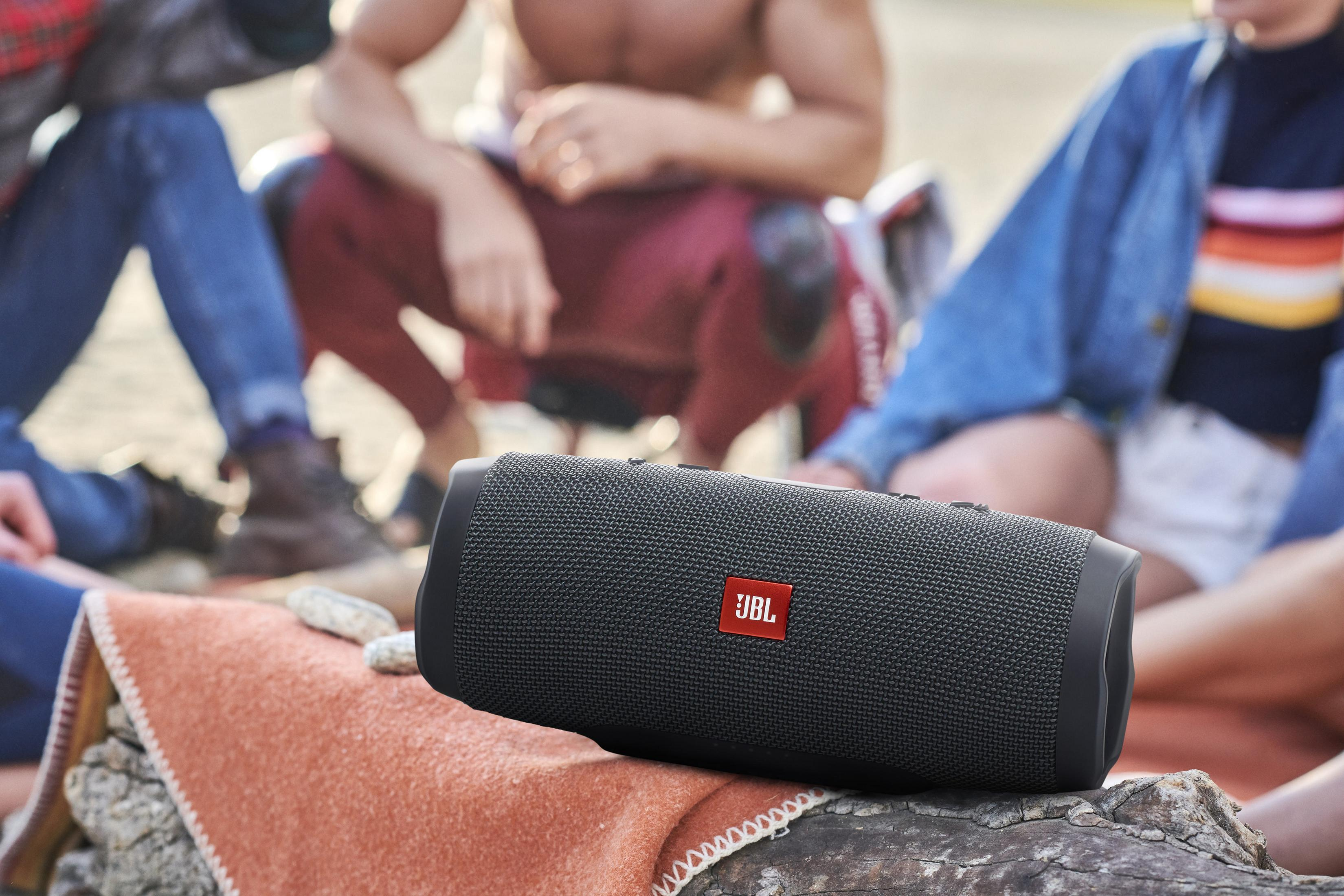 Altavoz JBL negro sobre un tronco. Gente sentada cerca, en una manta. Entorno soleado al aire libre.