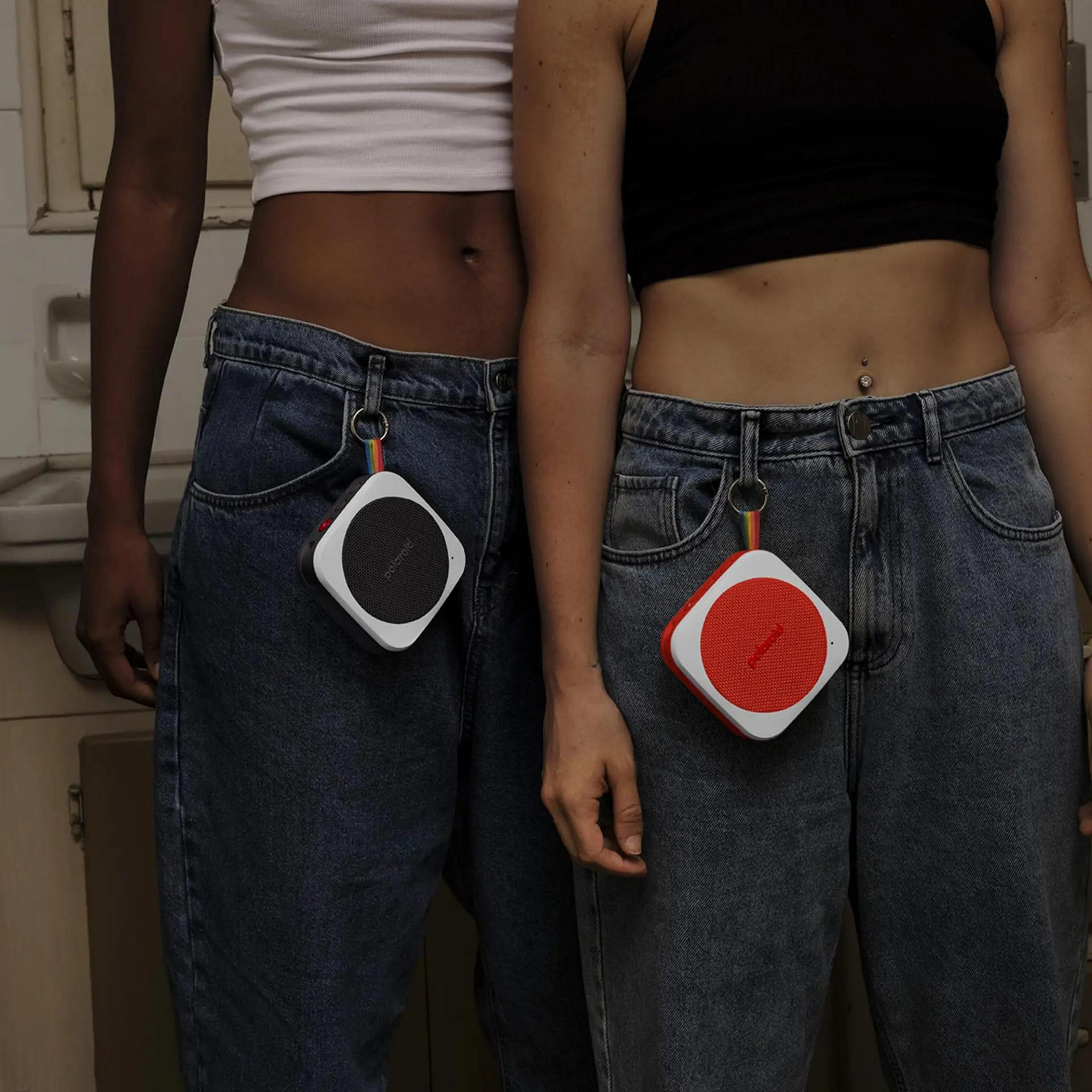 Dwie osoby z głośnikami wiszącymi z dżinsów. Jeden głośnik jest czerwony, drugi czarny. Białe tło.