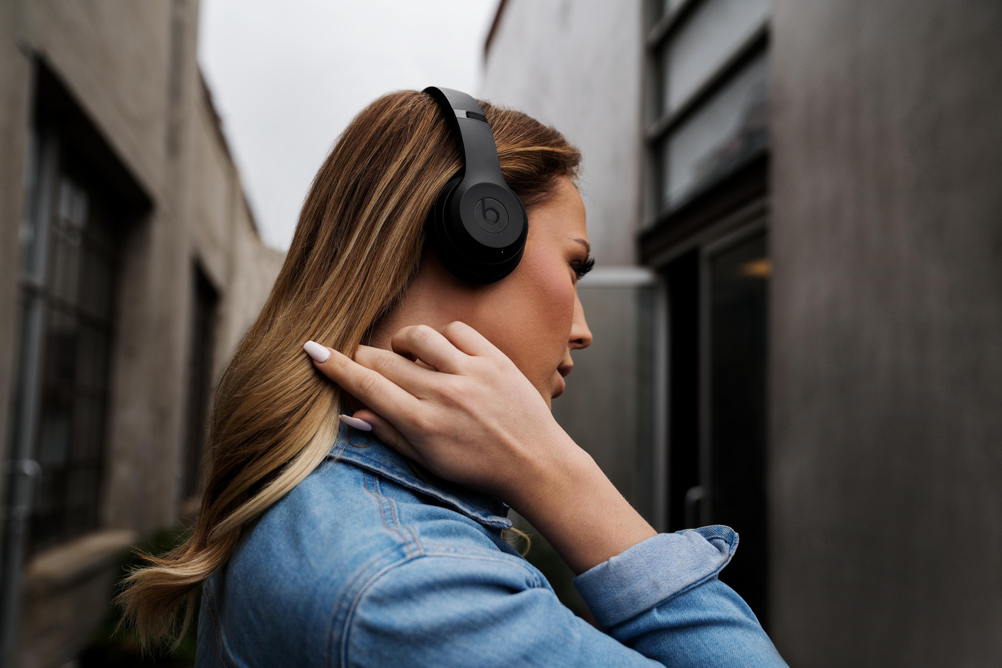 Mujer con cabello rubio usando auriculares negros y una chaqueta de mezclilla, en un callejón.