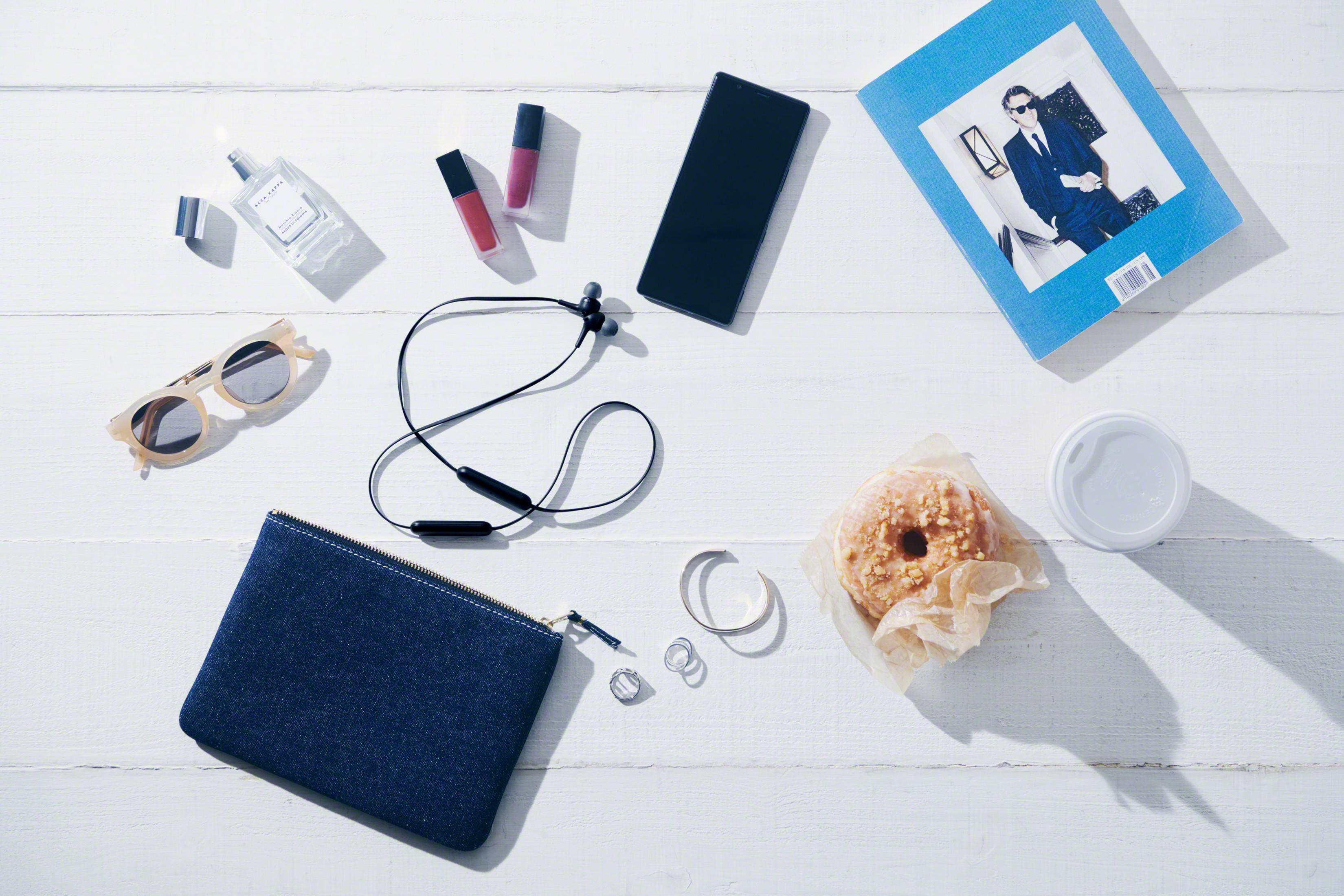 Ujęcie z góry na przedmioty codziennego użytku: okulary przeciwsłoneczne, perfumy, telefon, słuchawki, pączek i torba.