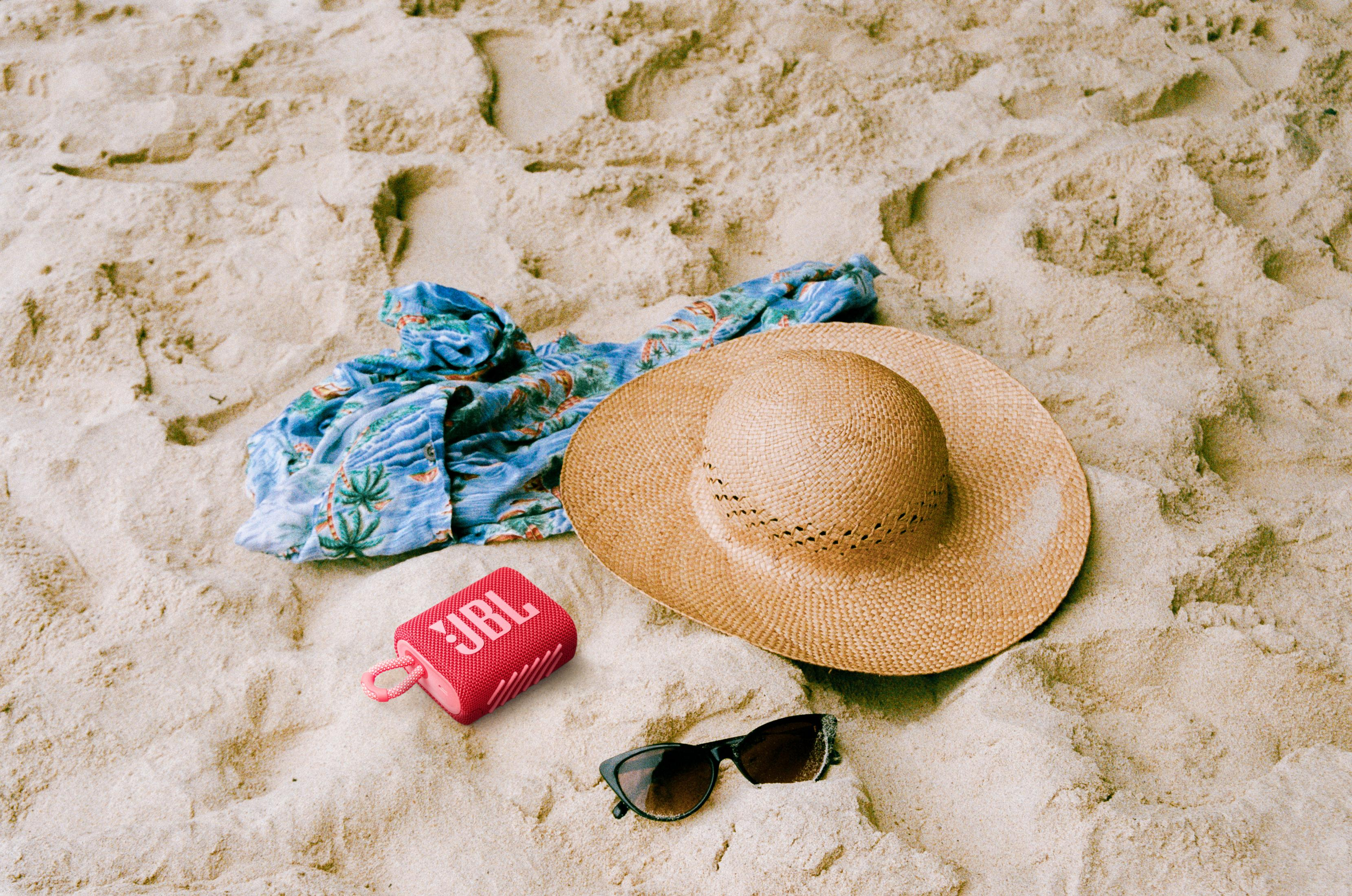 Scena na plaży: kapelusz, okulary przeciwsłoneczne, koszula i czerwony głośnik JBL na piasku.