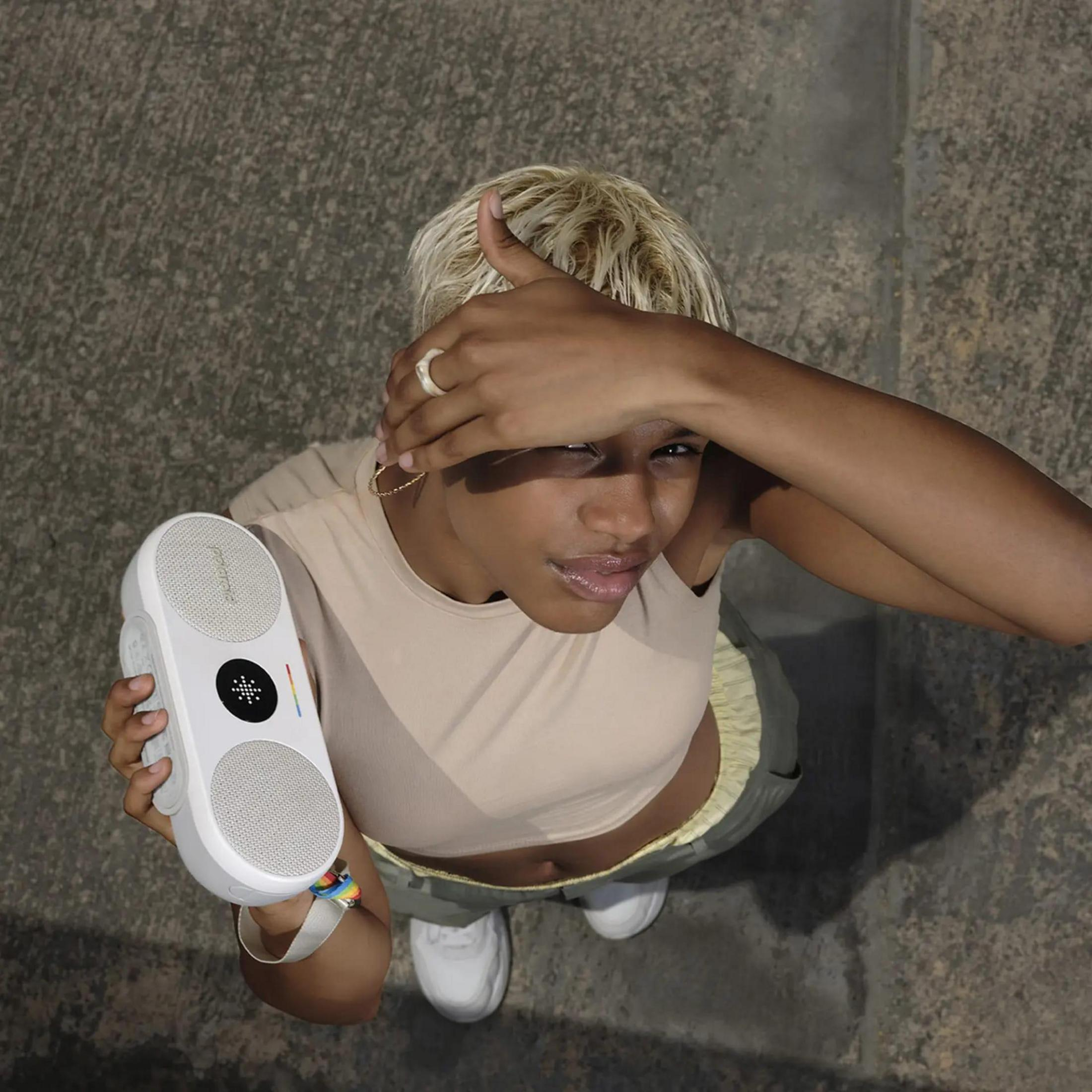 Kobieta trzymająca biały głośnik, zasłaniająca oczy dłonią, patrząca w górę.