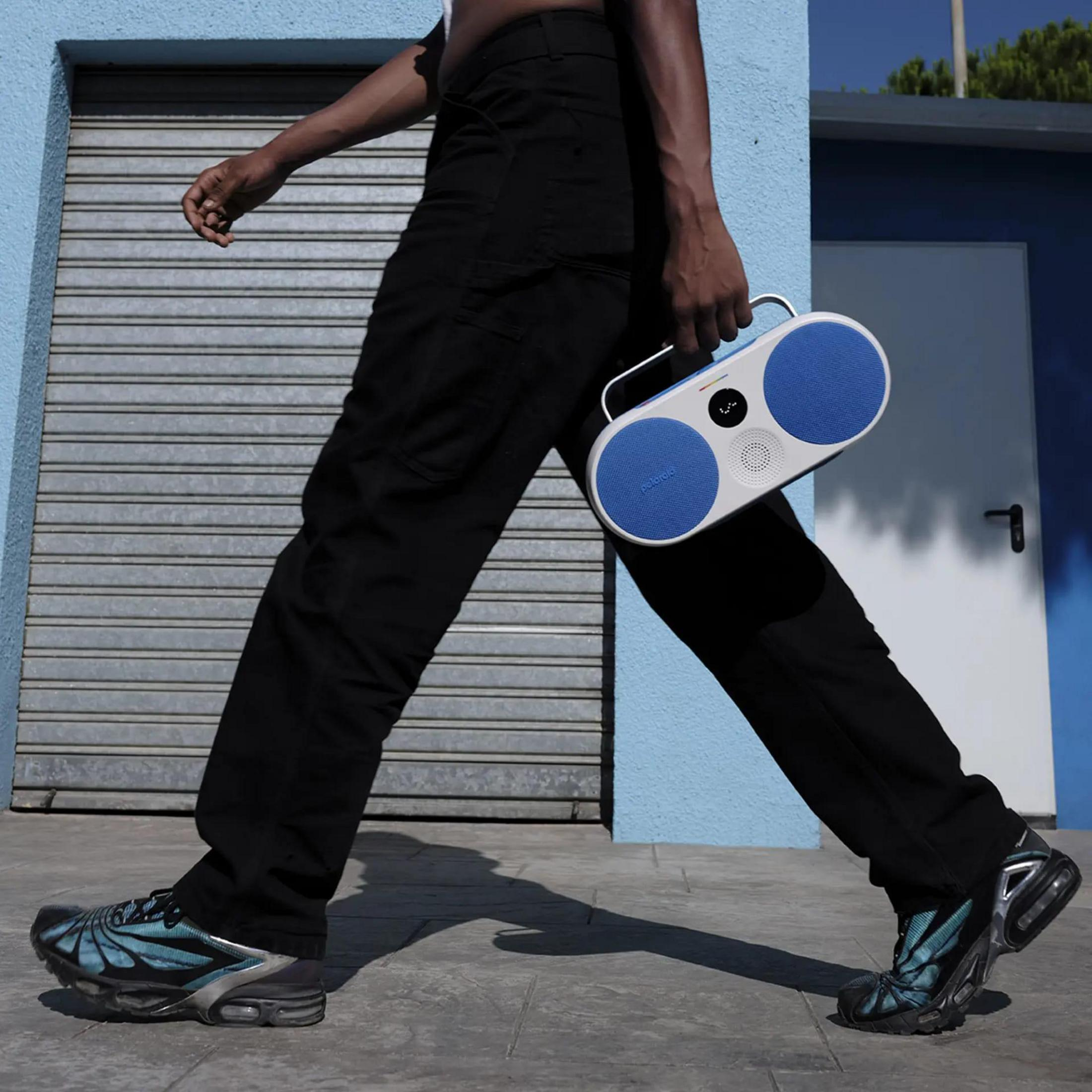 Osoba idzie z głośnikiem JBL. Niebiesko-biały głośnik, niebieska ściana. Nowoczesne otoczenie uliczne.