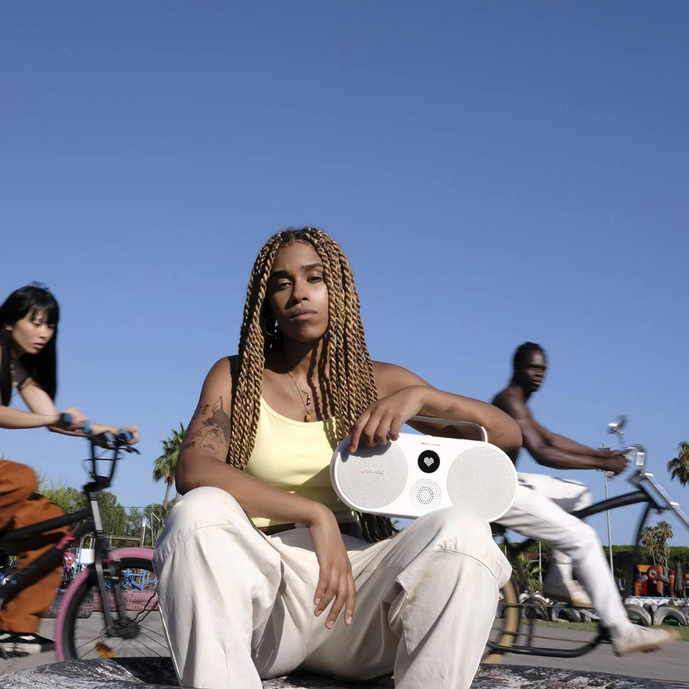 Kobieta trzyma biały głośnik. Dwie osoby na rowerach w tle, niebieskie niebo.