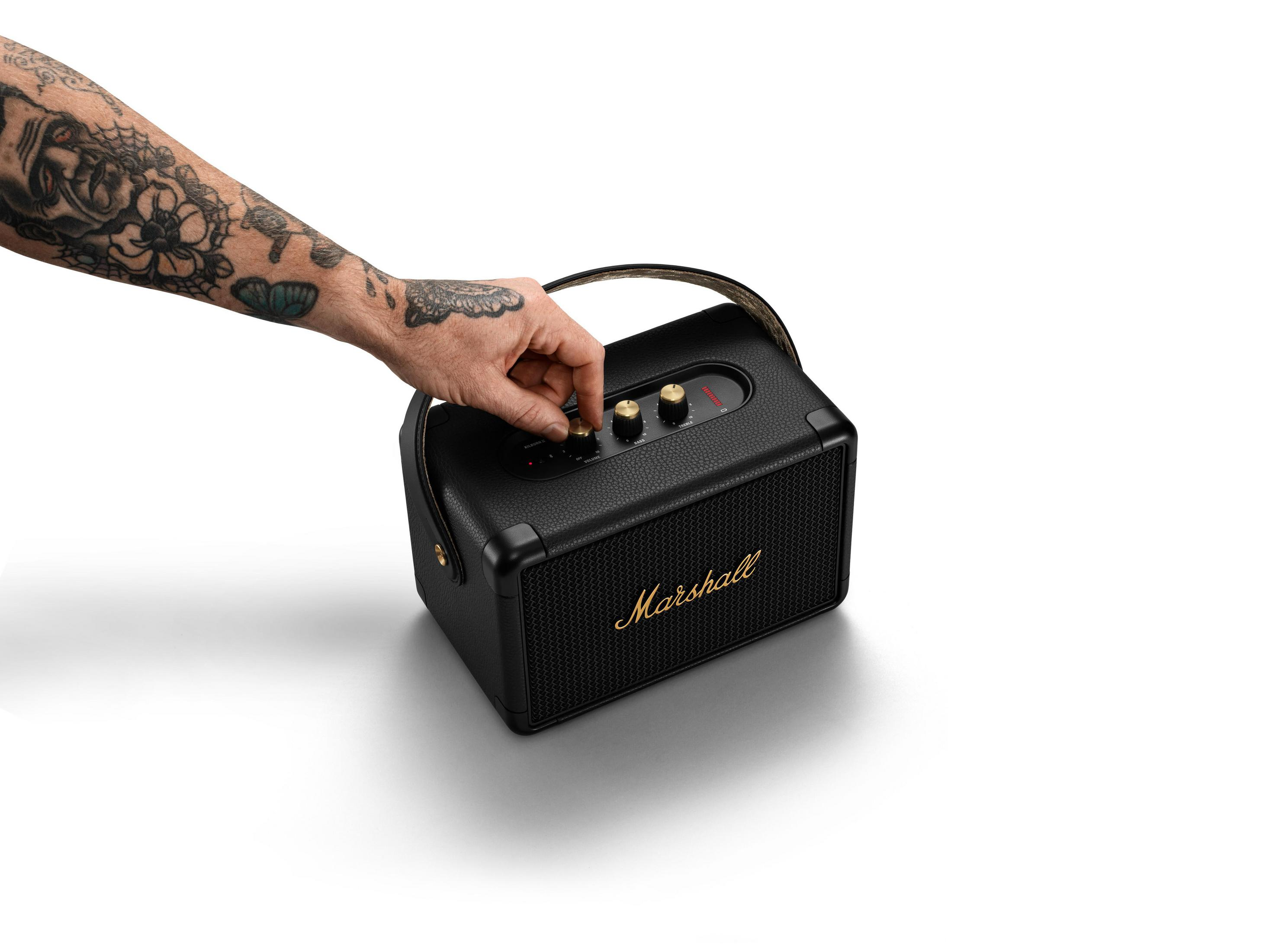 Eine Hand justiert den Knopf eines Marshall-Lautsprechers. Schwarzer Lautsprecher mit goldenen Akzenten. Weißer Hintergrund.