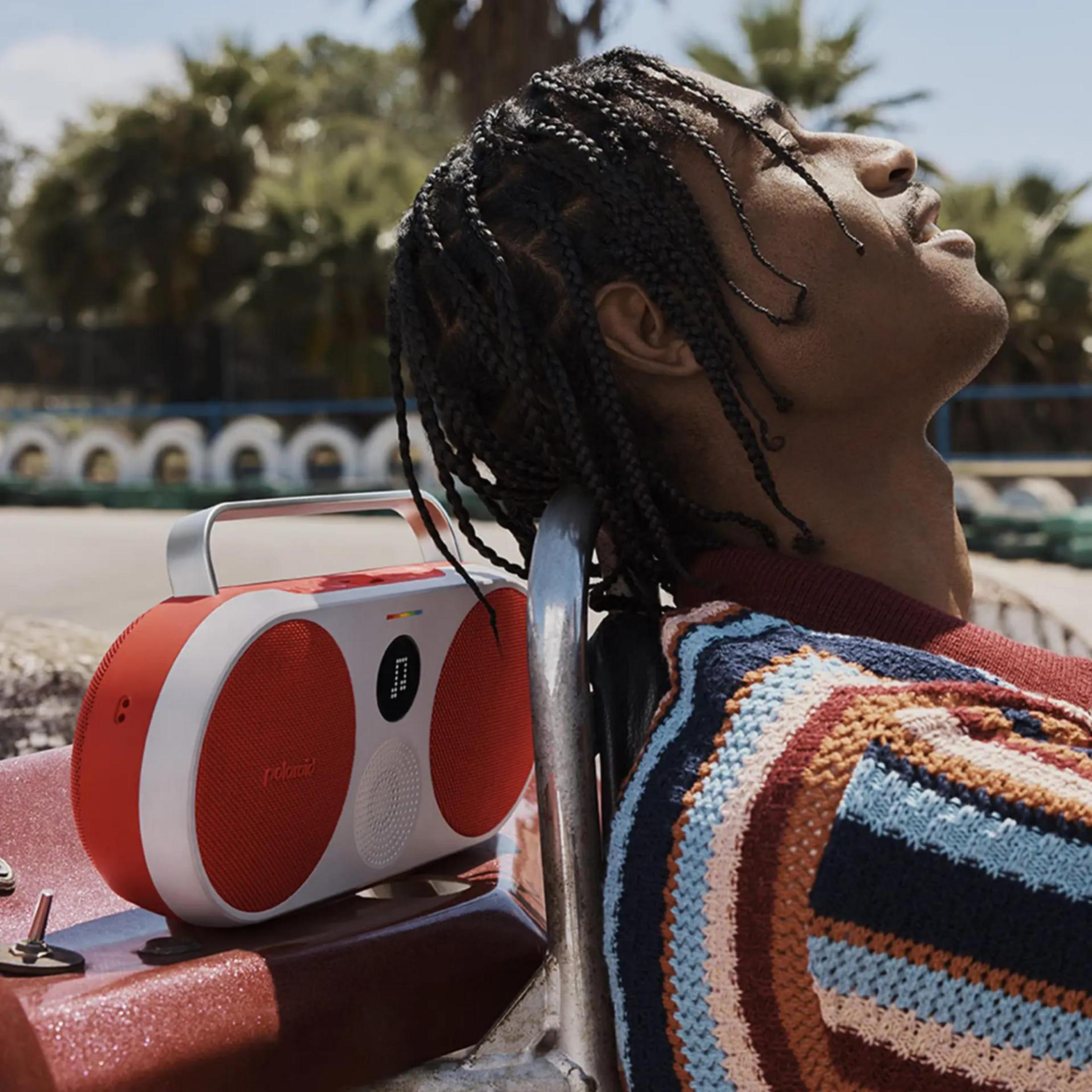 Mężczyzna z warkoczami leży obok pomarańczowo-białego boomboxa. Patrzy w górę.