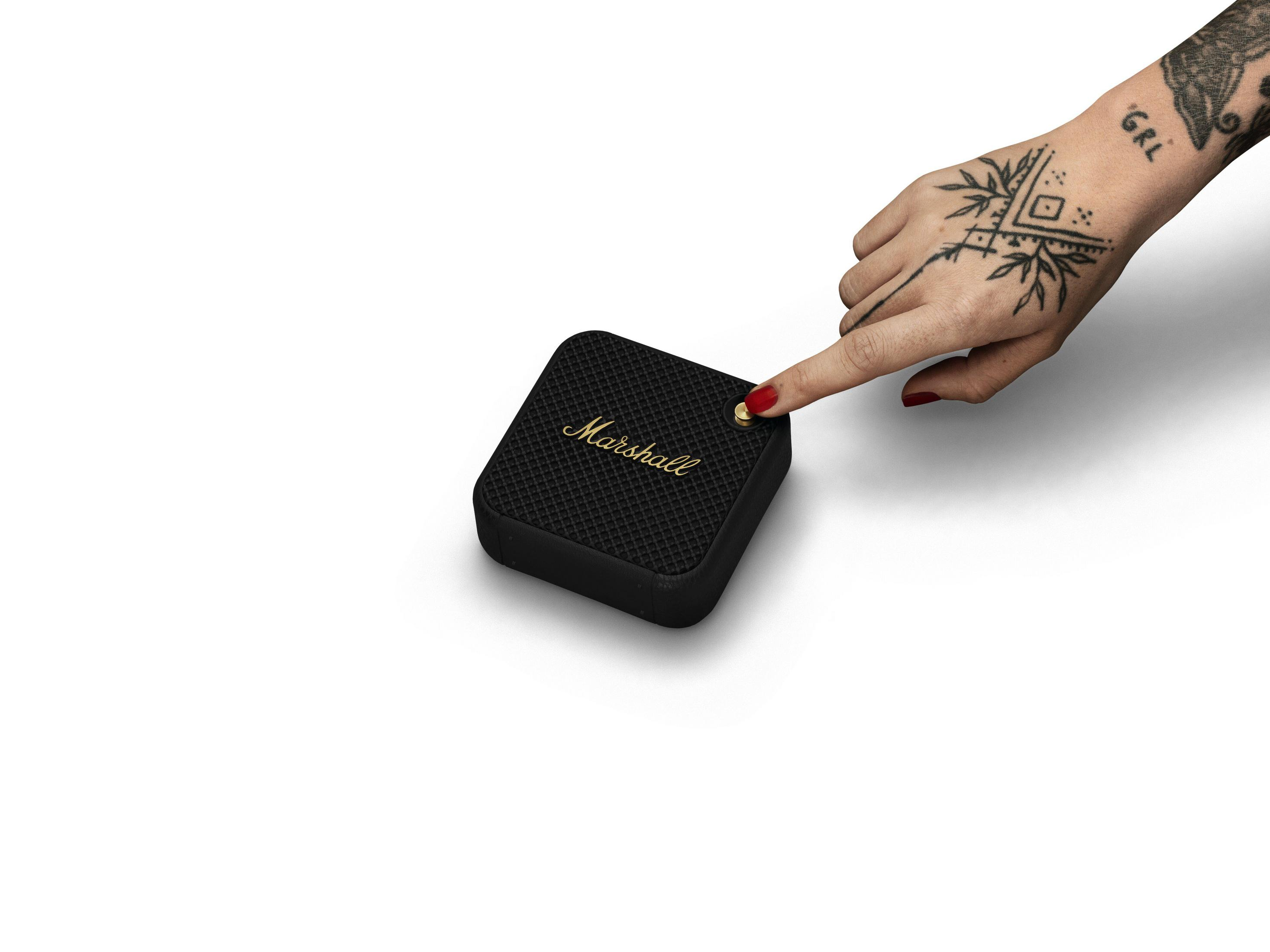 Una mano tatuata preme un pulsante su un altoparlante Marshall nero su sfondo bianco.