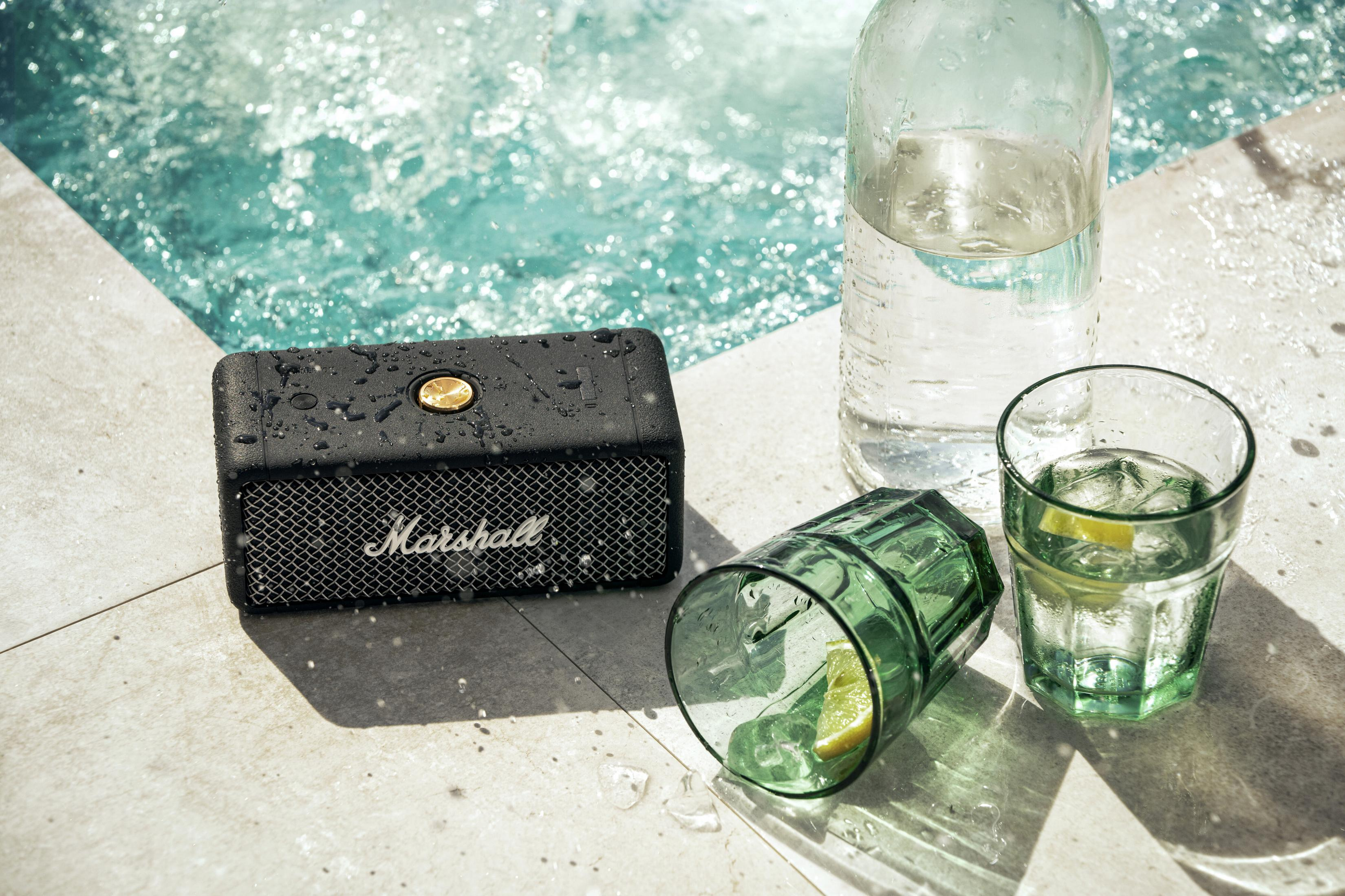 Ein schwarzer Marshall-Lautsprecher befindet sich in der Nähe eines Pools. Ein Glas Wasser und eine Flasche sind auch da.