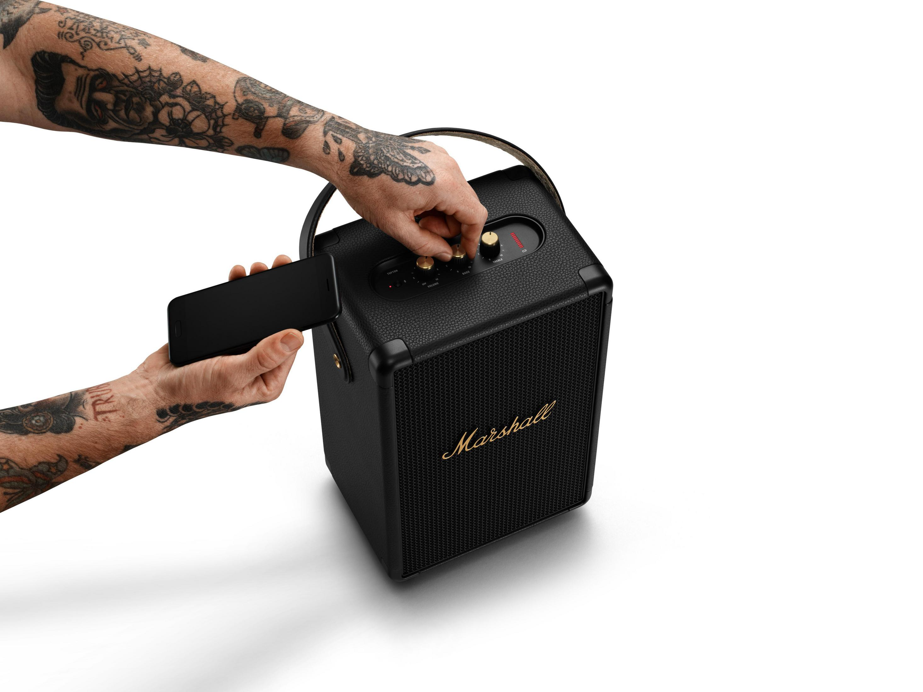 Hombre con tatuajes ajusta el volumen de un altavoz Marshall negro, sosteniendo un teléfono.