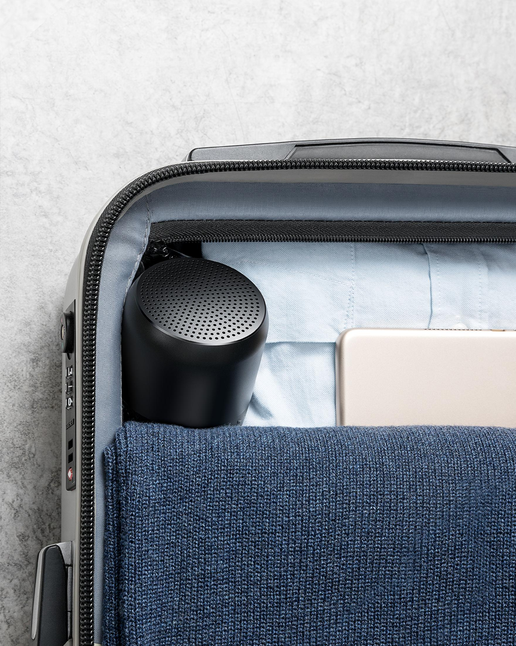 Czarny głośnik w szarej walizce. Głośnik znajduje się obok tabletu i niebieskiego swetra.