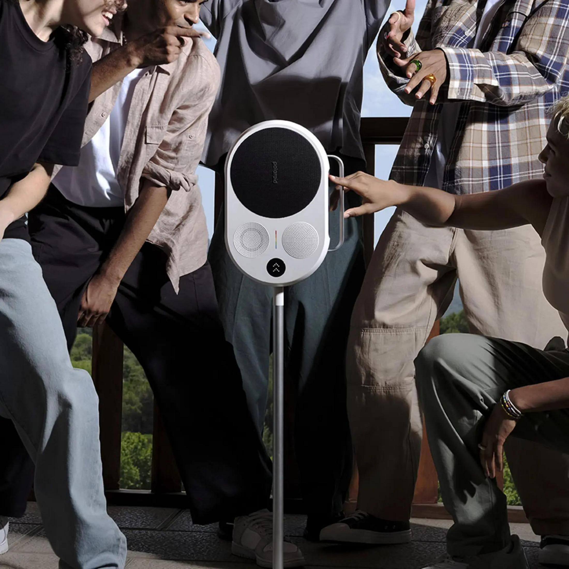 Biały głośnik Polaroid na stojaku, otoczony przez grupę ludzi.