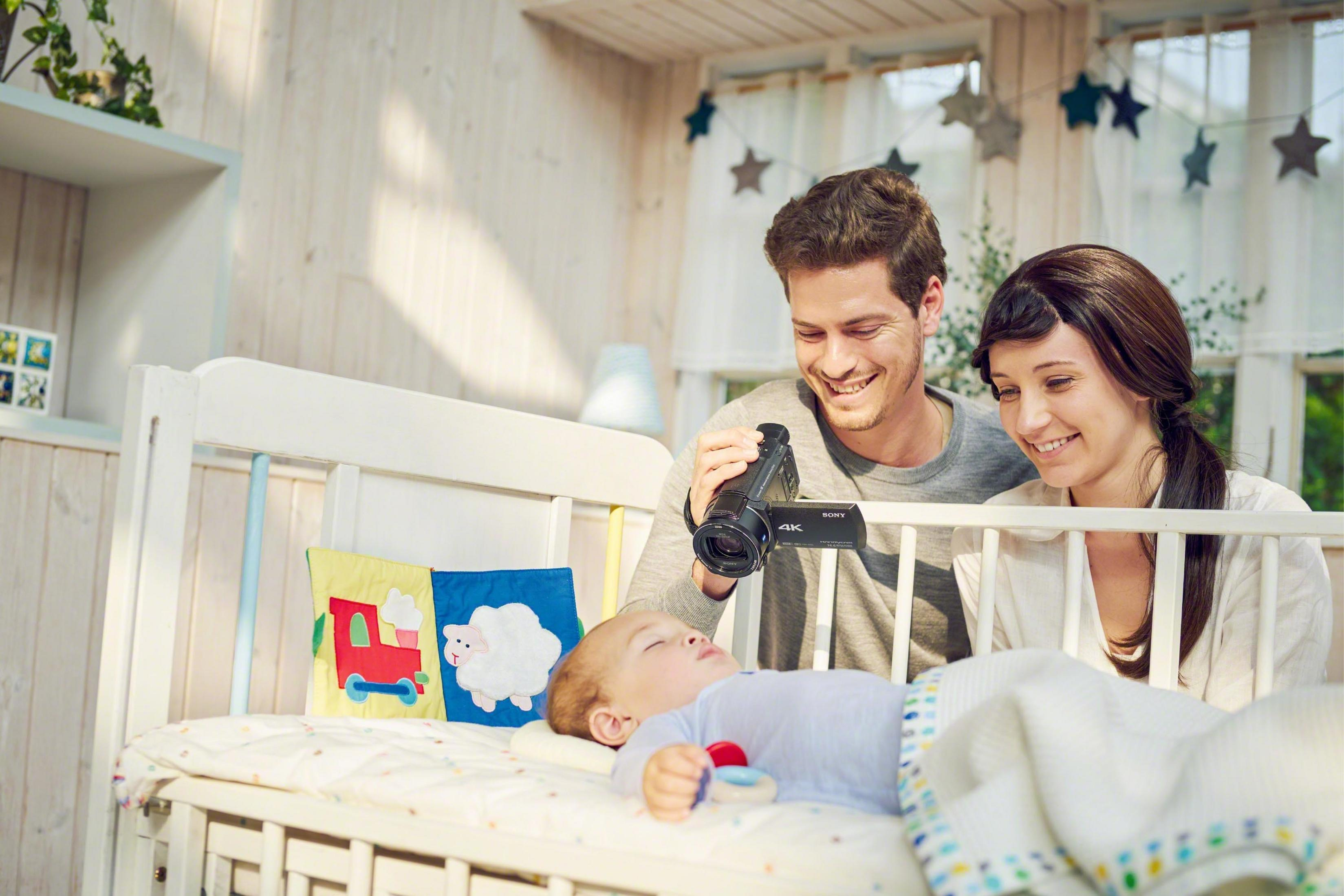 Eltern filmen ein Baby in einem Kinderbett, lächelnd, in einem dekorierten Raum.