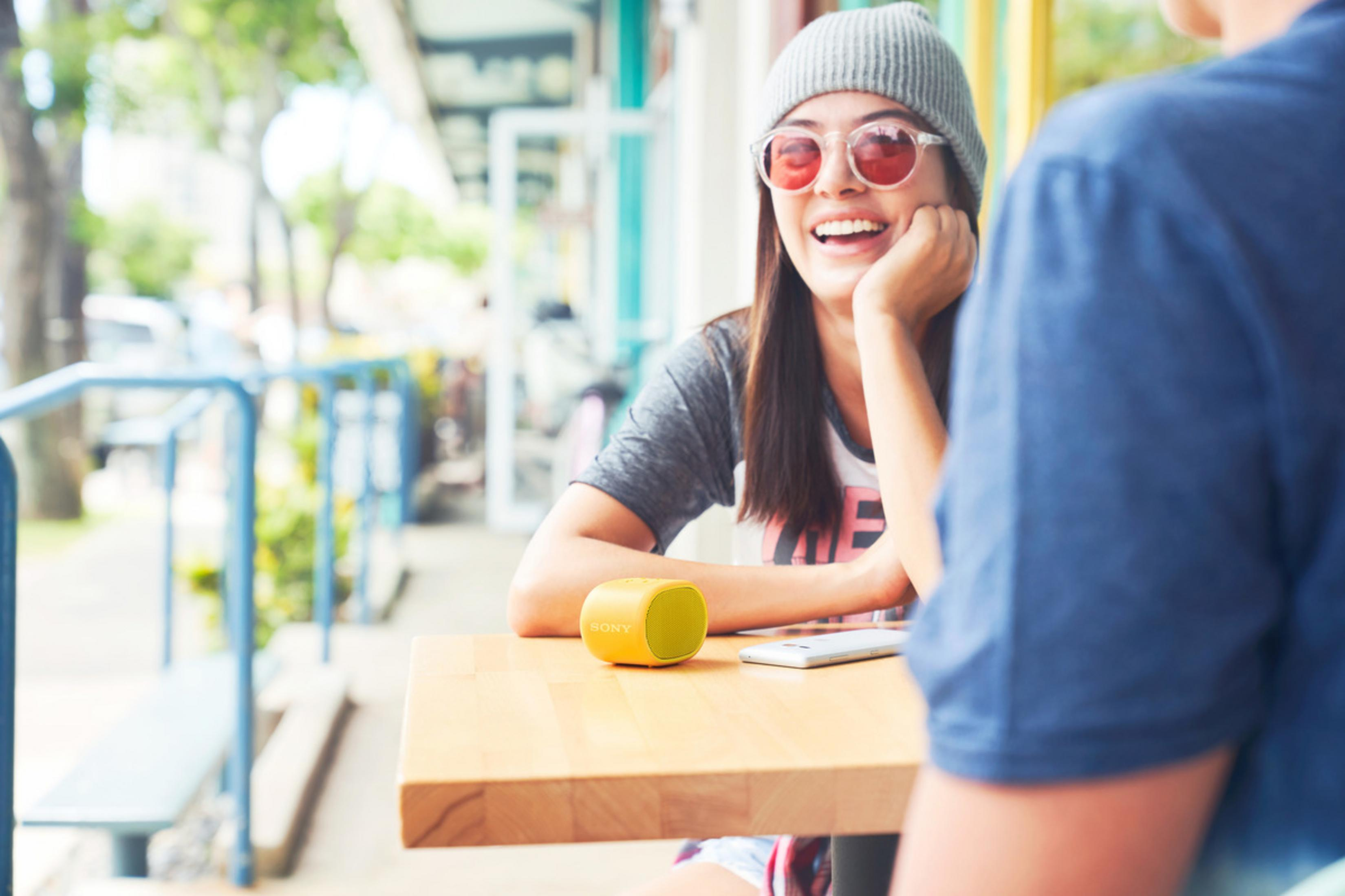 Mujer sonríe en una mesa de café con un altavoz amarillo. Otra persona es parcialmente visible.