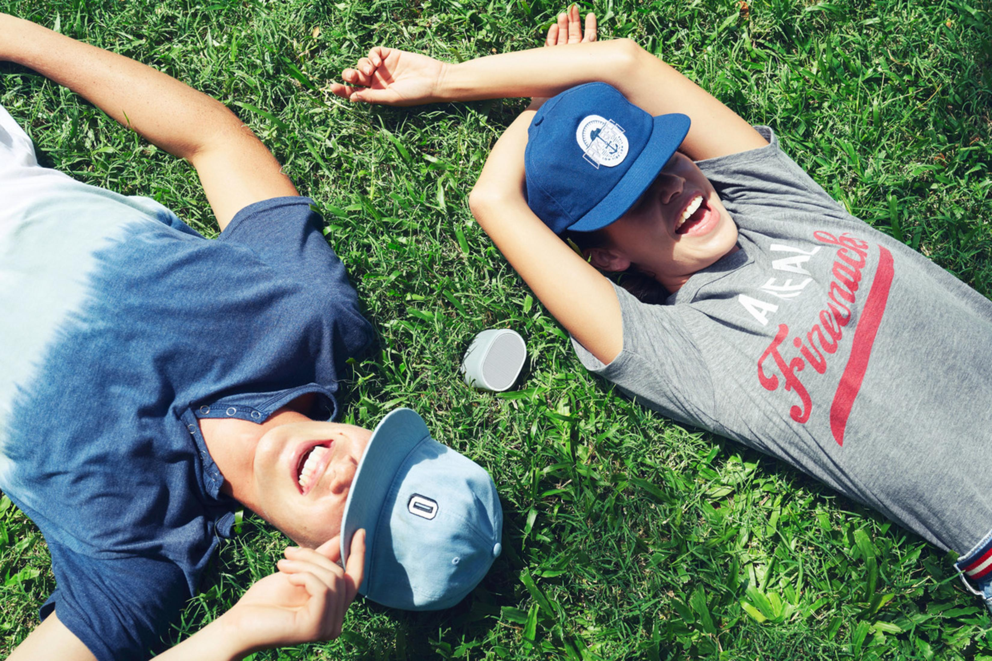 Dwie osoby śmiejące się na zielonej trawie w czapkach i z małym głośnikiem.