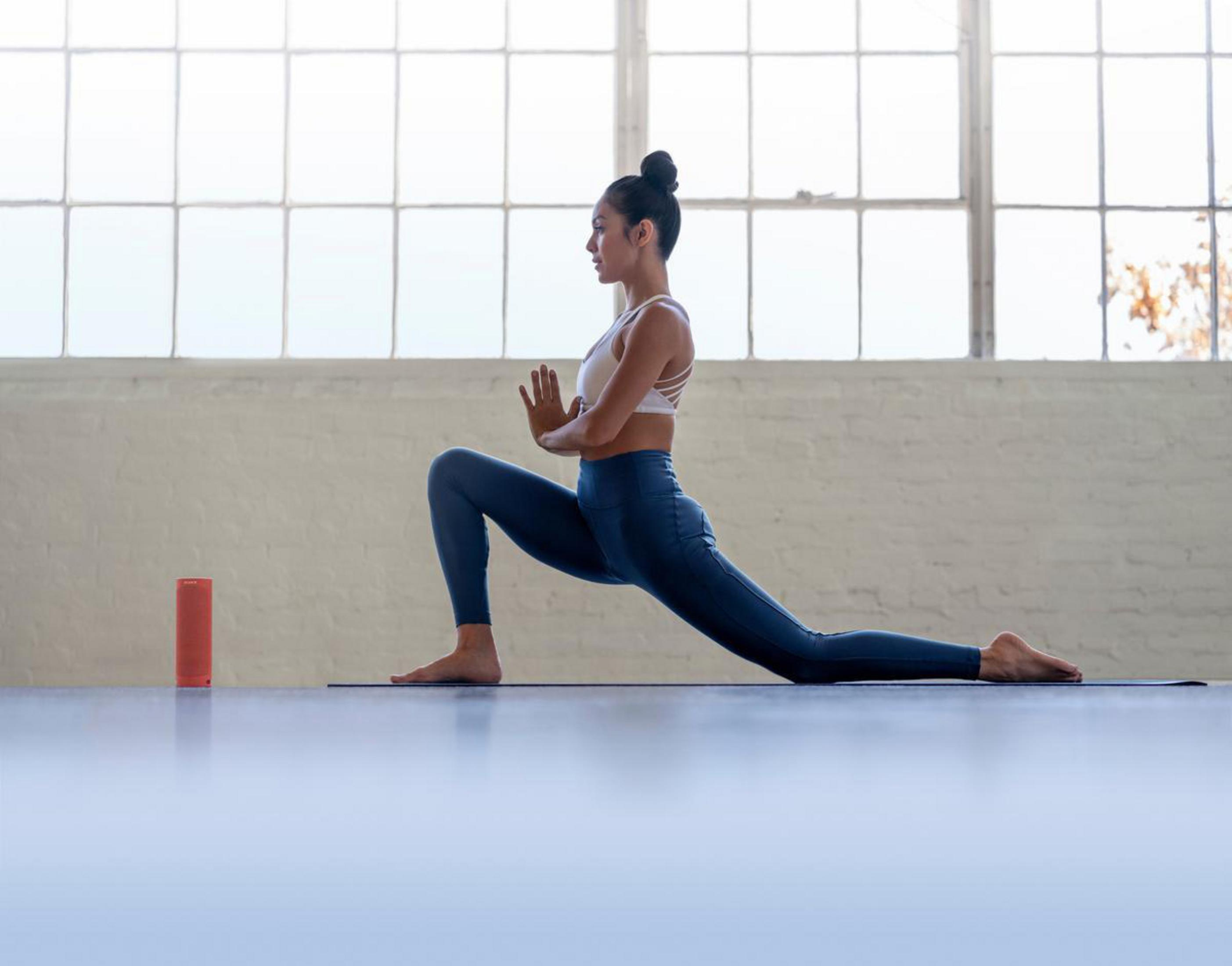 Frau macht Yoga in einem Fitnessstudio. Sie ist in einer Ausfallschrittstellung. Es gibt einen Lautsprecher und ein Fenster.