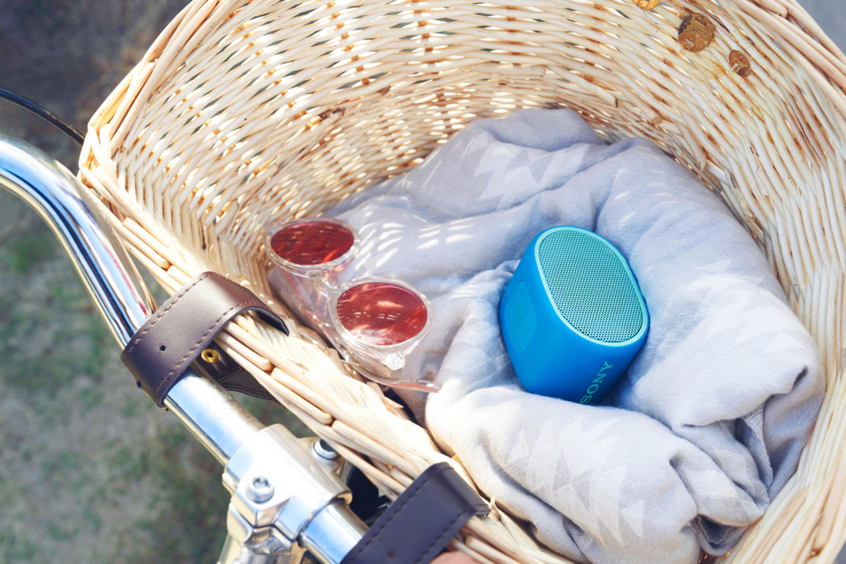 Cesta de mimbre en el manillar de la bicicleta, gafas de sol, altavoz azul y tela gris.